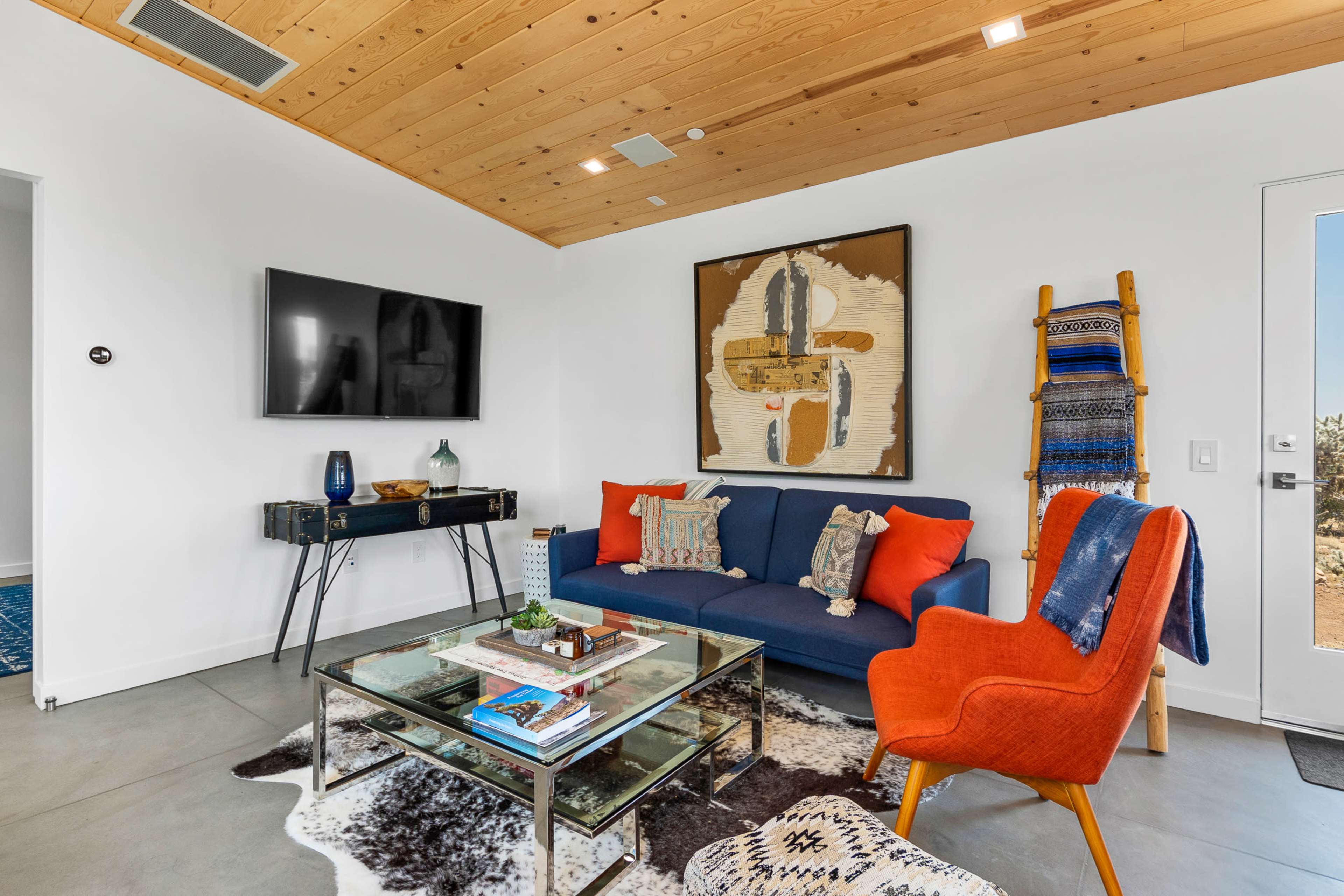 A modern living room featuring a blue sofa, an orange armchair, a glass coffee table, and a large wall-mounted television.