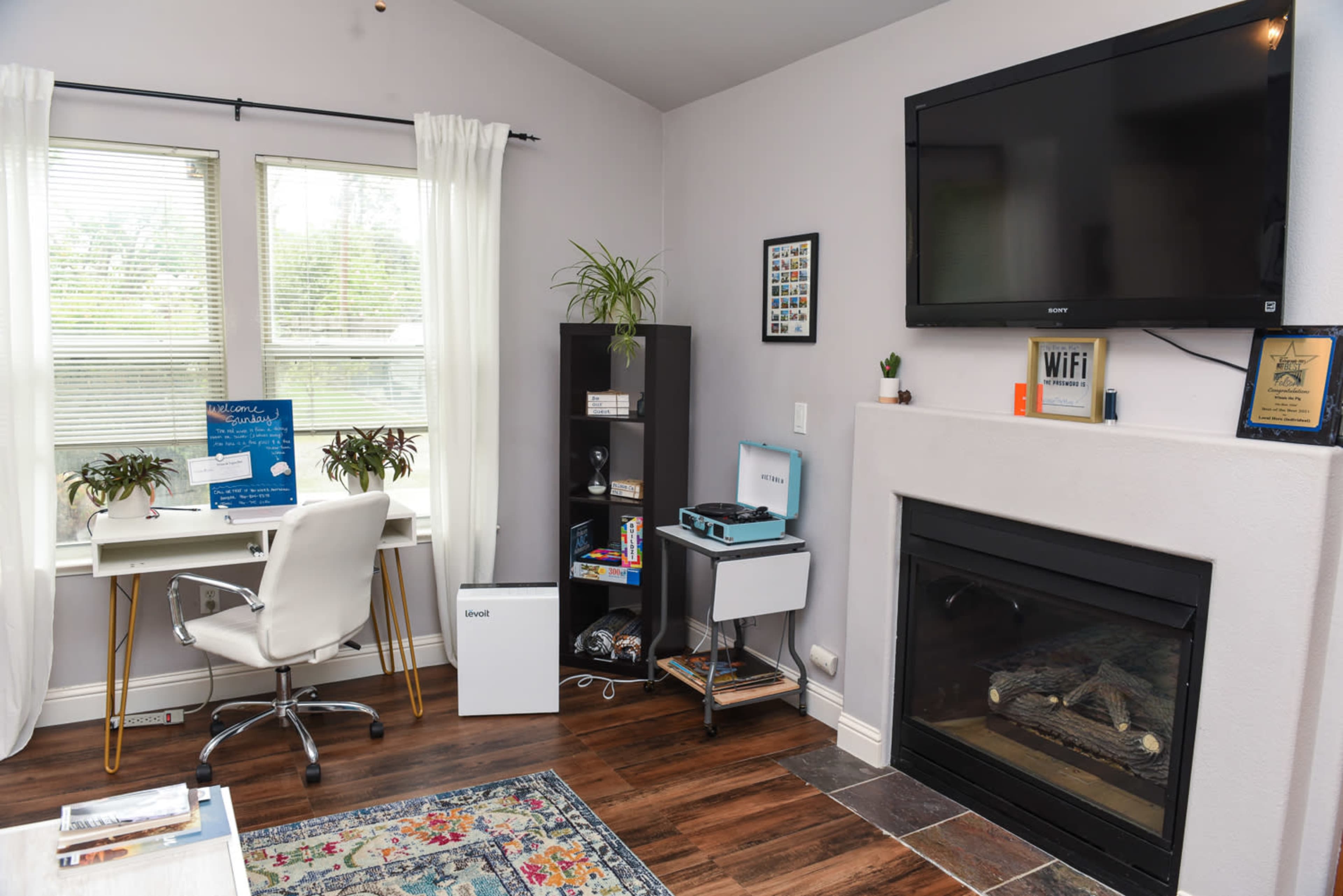 The image shows a home office setup featuring a white desk with a chair, a TV mounted on the wall, a bookshelf, and a fireplace in a light-colored room.
