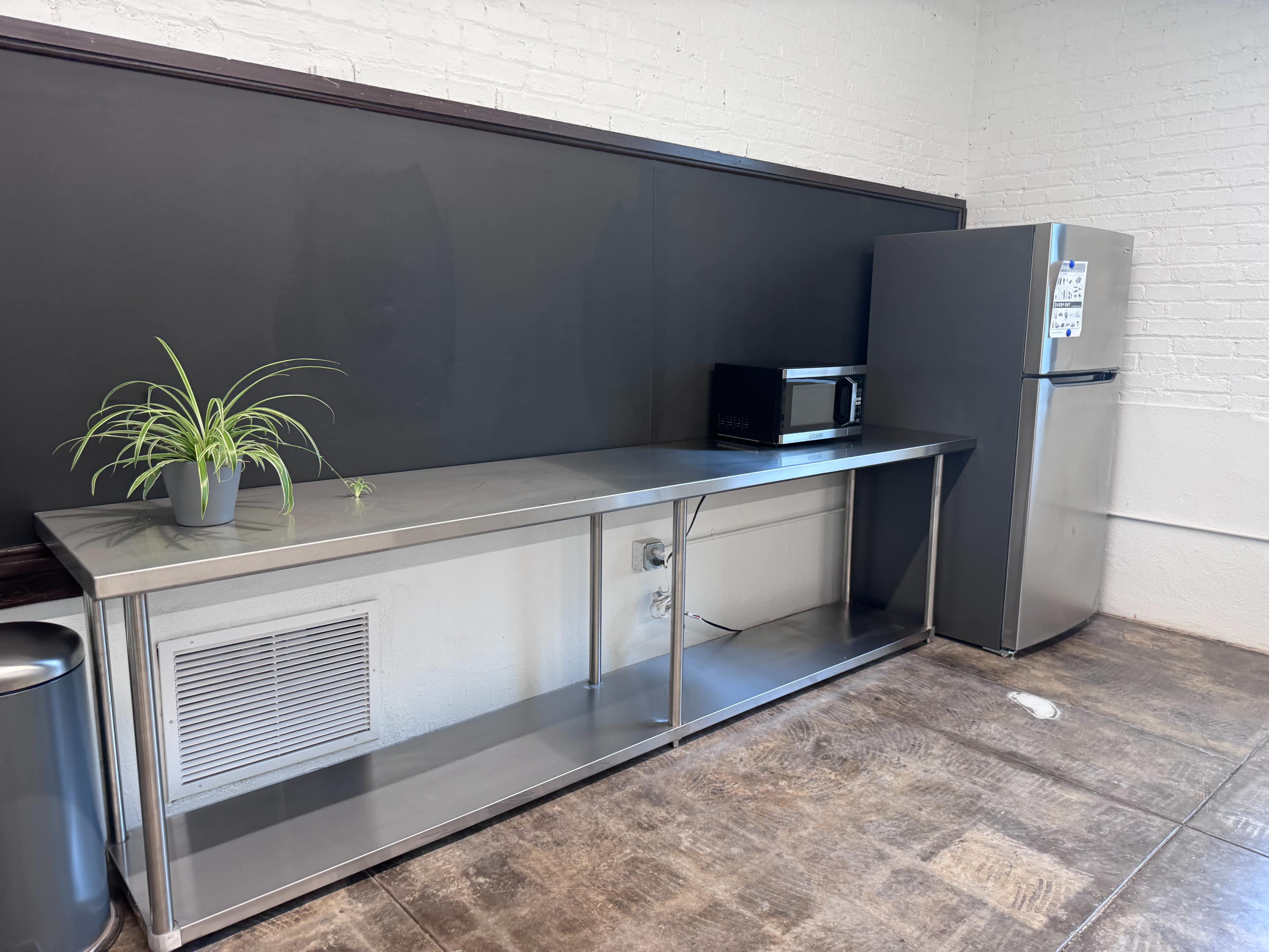 The image shows a kitchen area with a stainless steel table, a microwave, a small plant, a refrigerator, and a trash can against a white brick wall.