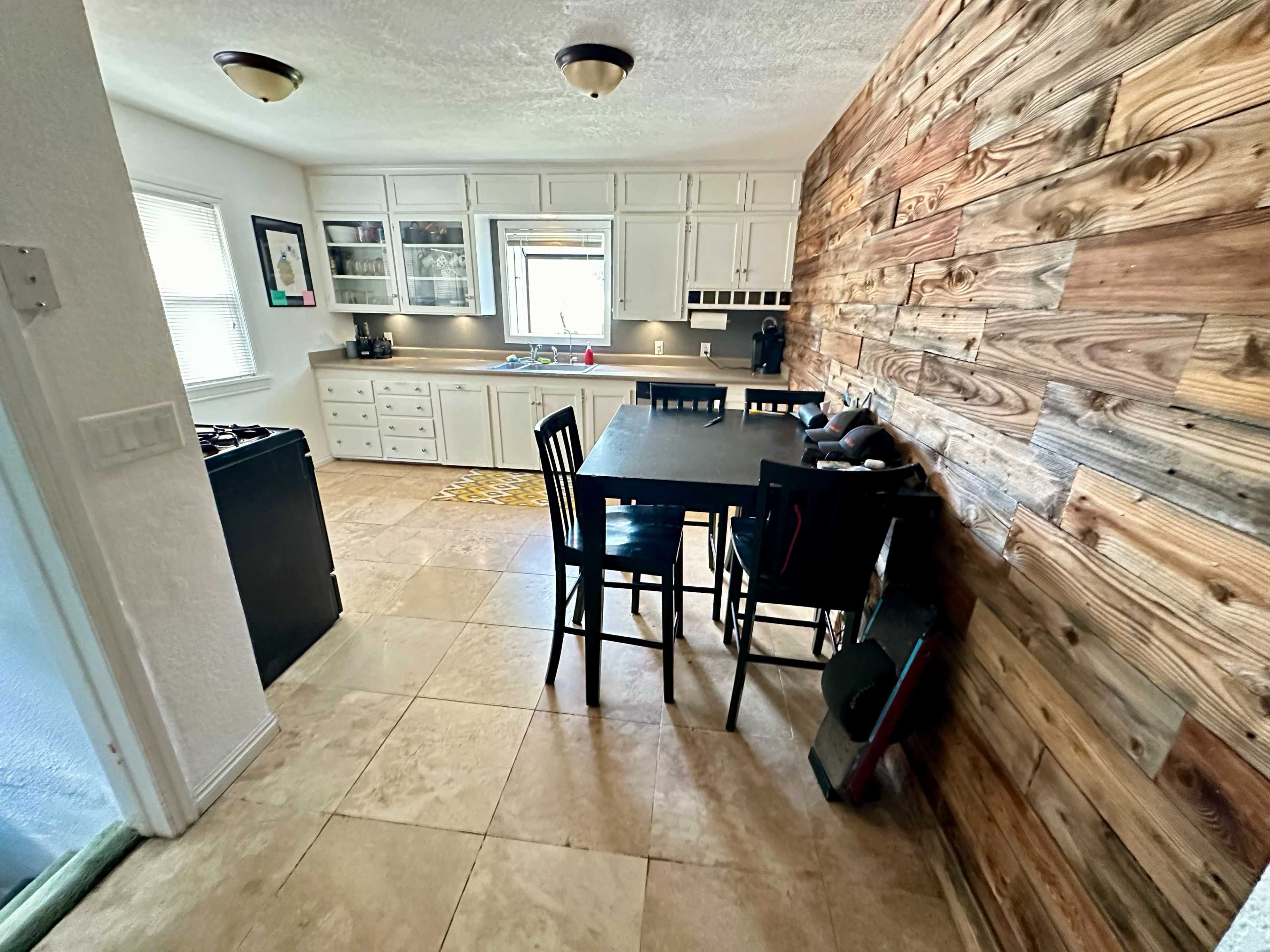 A kitchen features a wooden accent wall, a dining table with black chairs, and white cabinetry against tiled flooring.