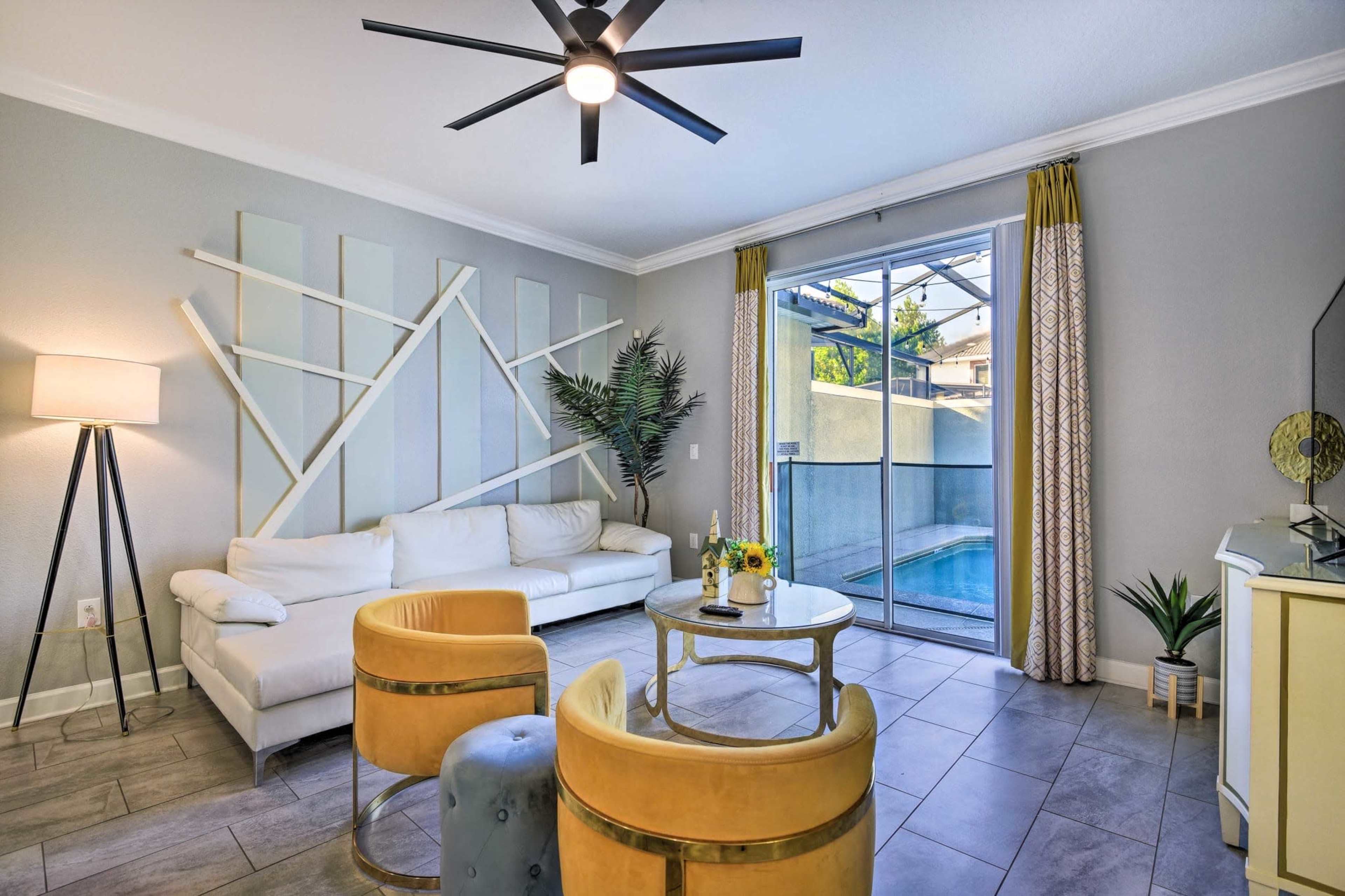 A modern living room features a white sectional sofa, yellow accent chairs, a round coffee table, and large windows overlooking a pool area.