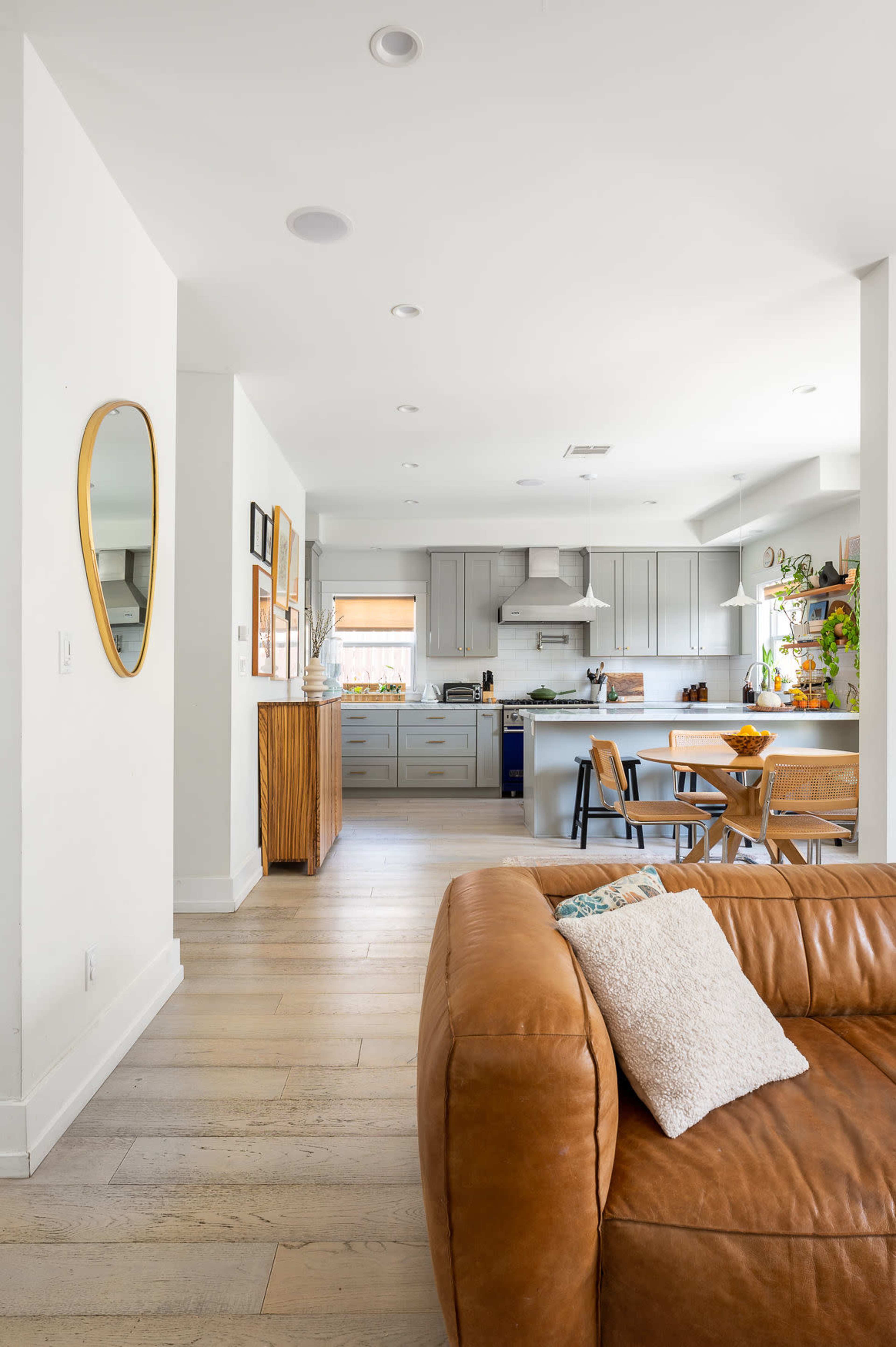 A modern kitchen and dining area, featuring light wood flooring, grey cabinetry, and a leather sofa in the foreground.