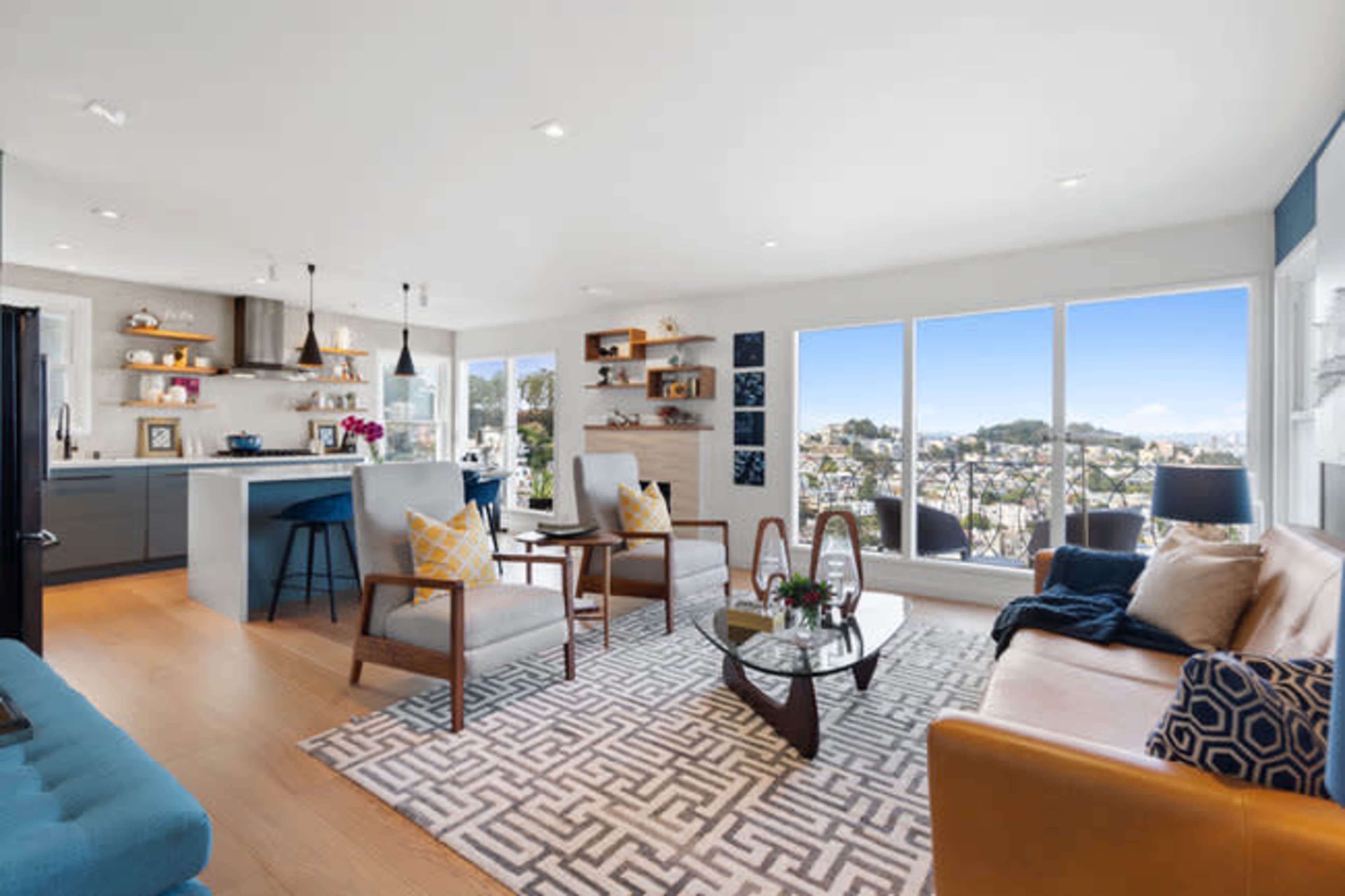 The image shows a modern living room featuring large windows with a view, a mix of furniture including a brown sofa and white chairs, and a geometric patterned rug.