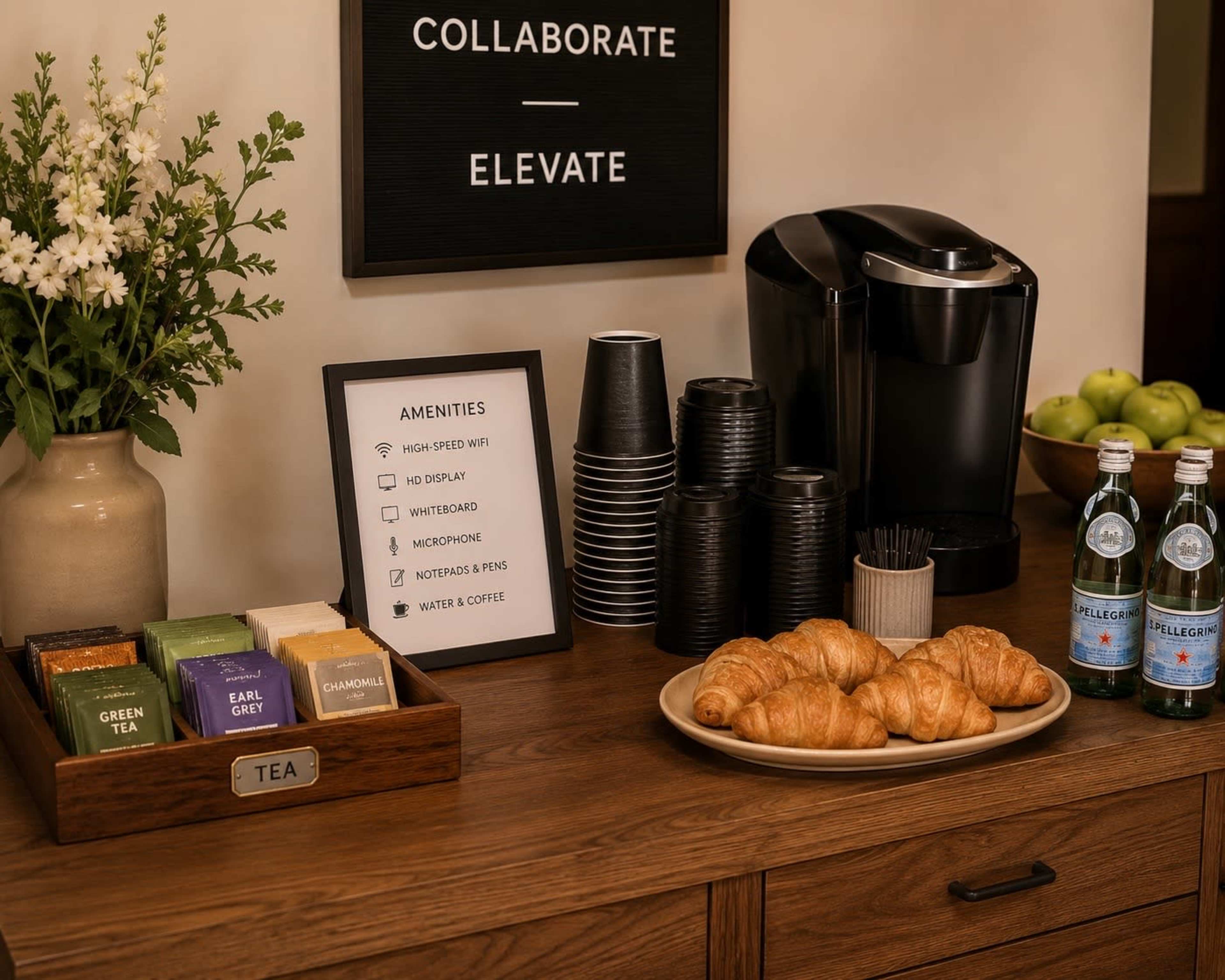 A coffee station is set up on a wooden table, featuring a coffee maker, tea selection, croissants, and bottled water.