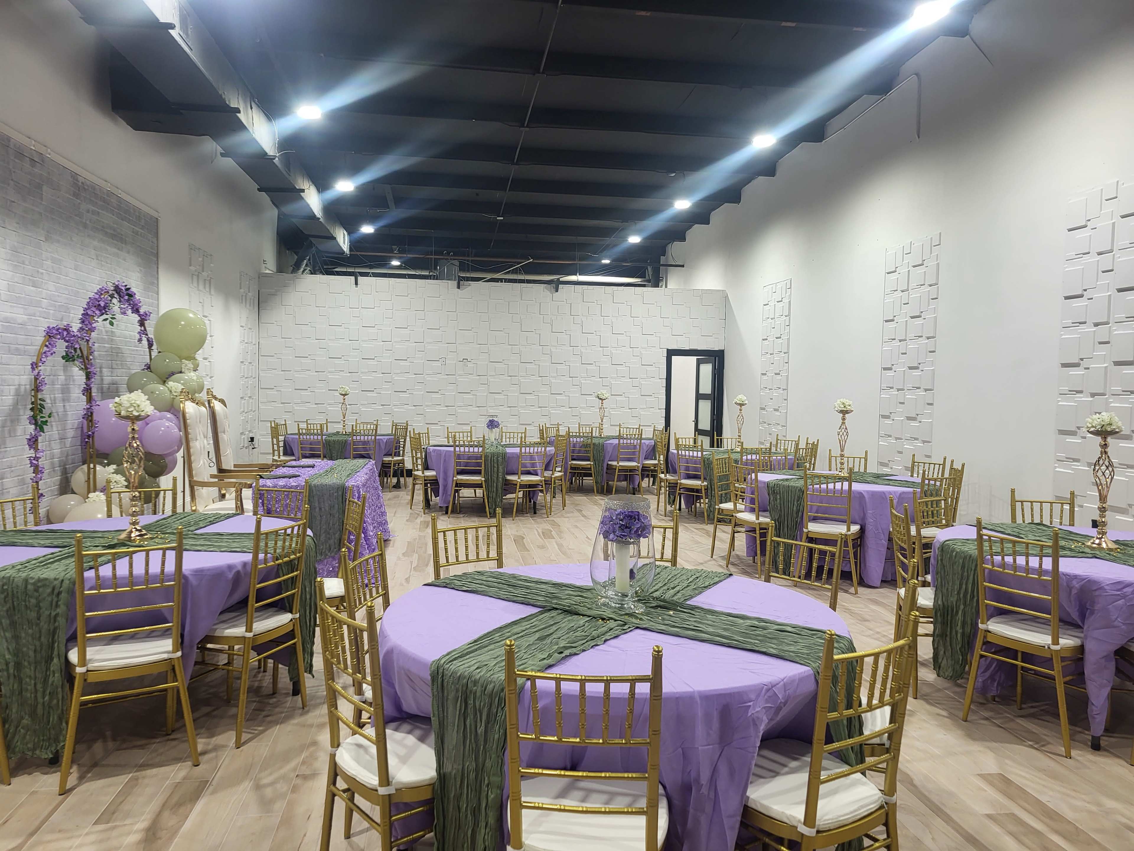 The image depicts a banquet hall set up for an event, featuring several round tables covered with purple and green tablecloths and surrounded by gold chairs.