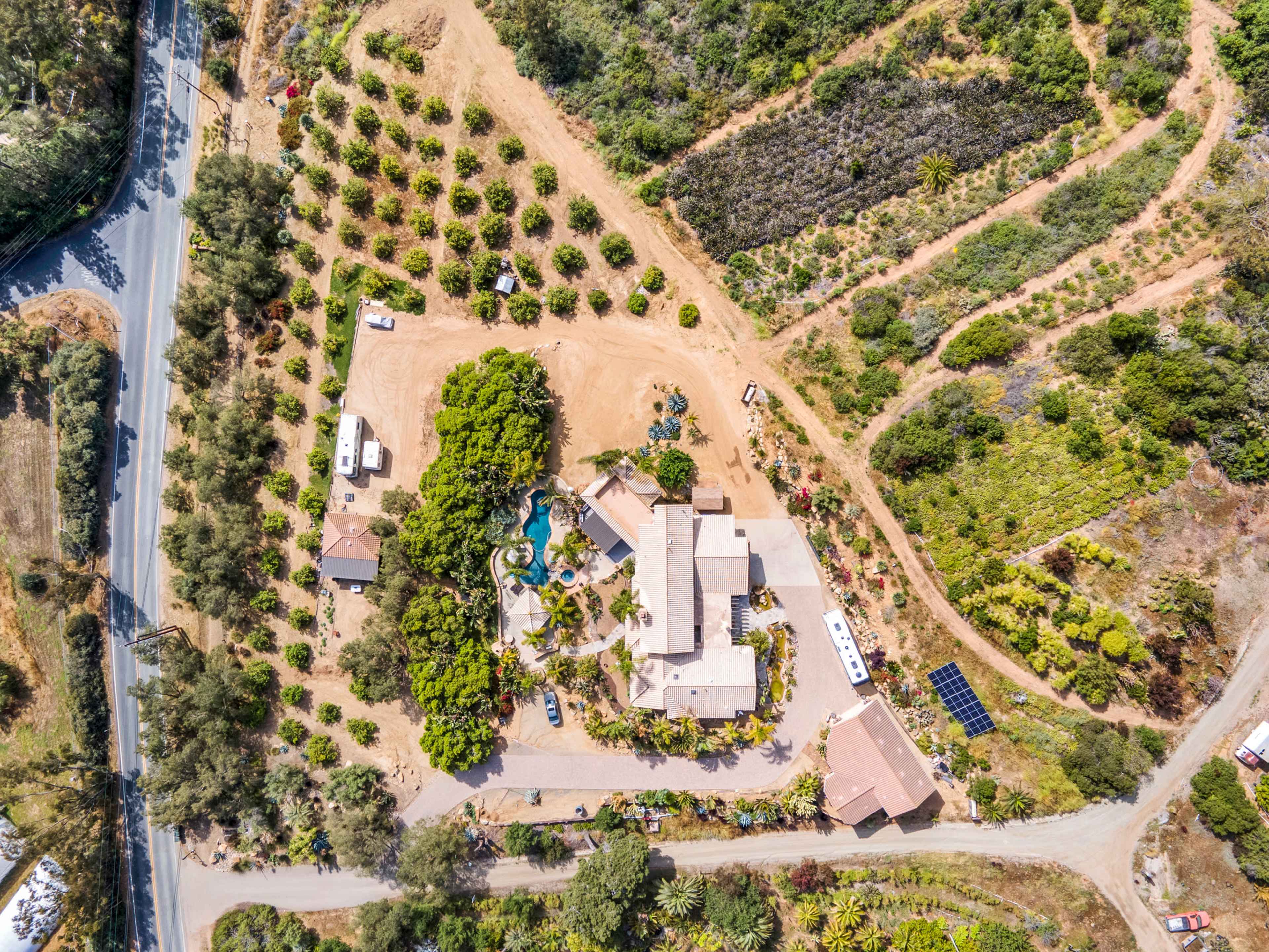 An aerial view of a large estate featuring a main house, landscaped gardens, a pool, and surrounded by fruit trees and greenery.