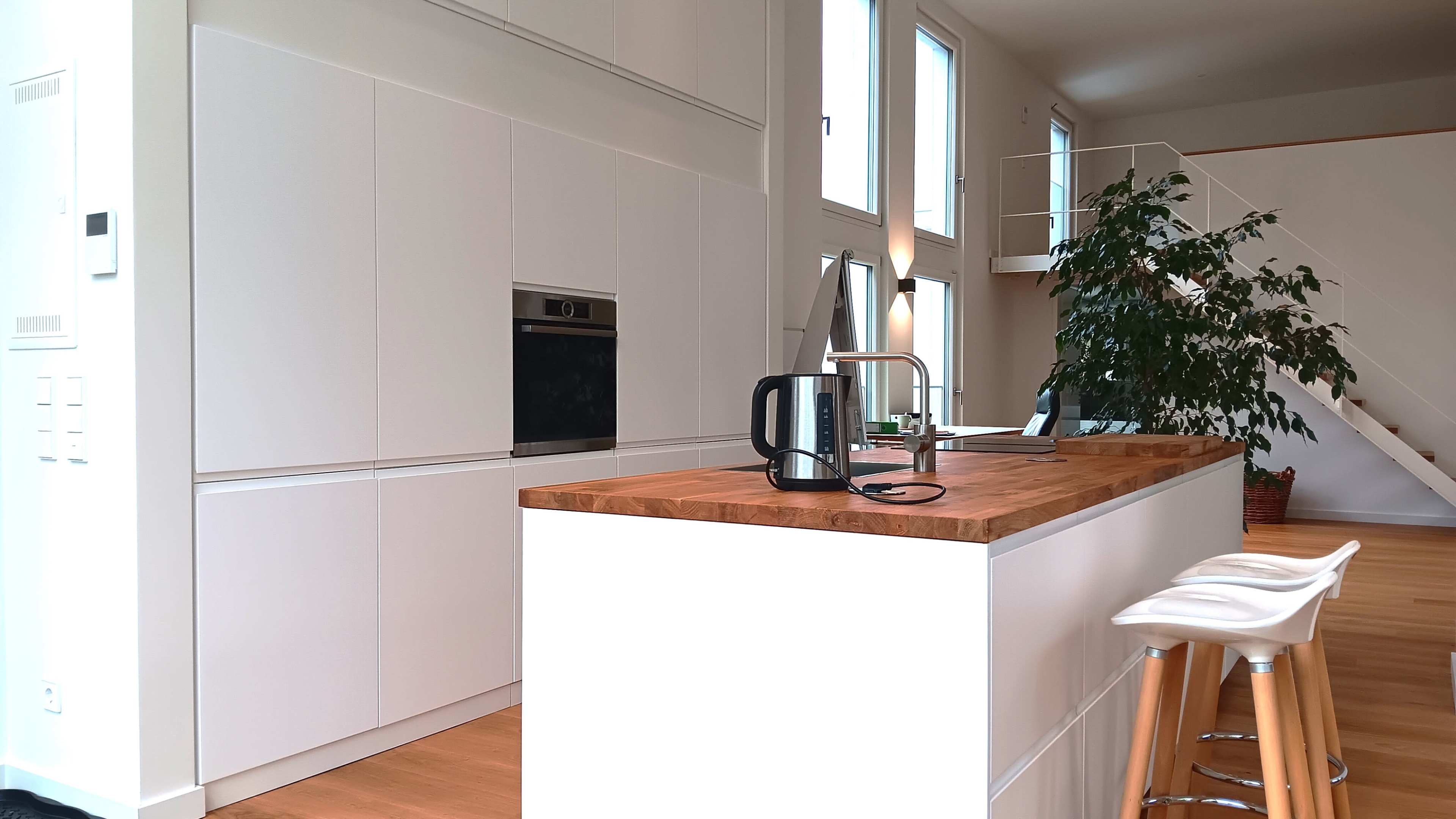 A bright kitchen with white cabinetry, a wooden countertop island, a kettle, and a staircase in the background.