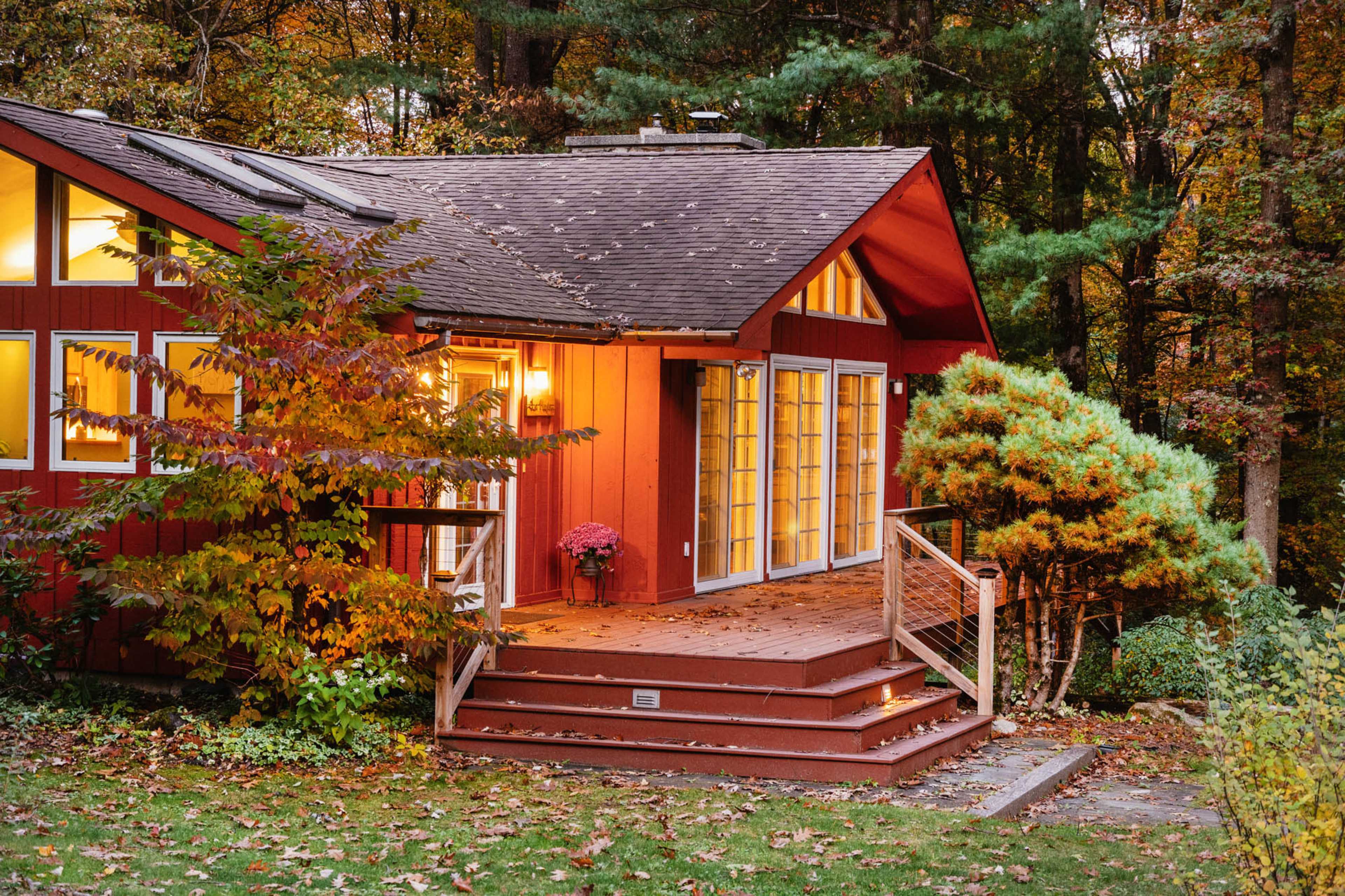 A red house with large windows and a wooden deck is surrounded by autumn foliage.