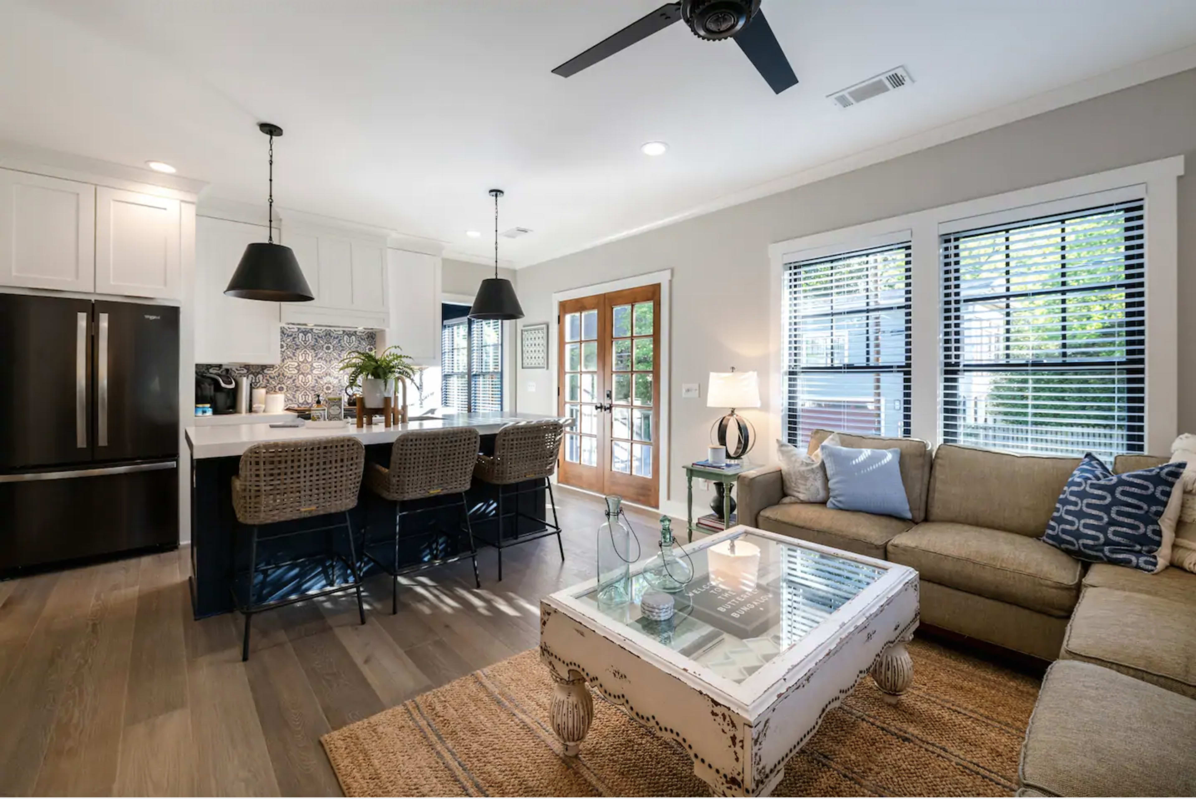 The image shows a modern living area featuring a comfortable sofa, a coffee table, a kitchen with bar seating, and large windows that allow natural light to fill the space.