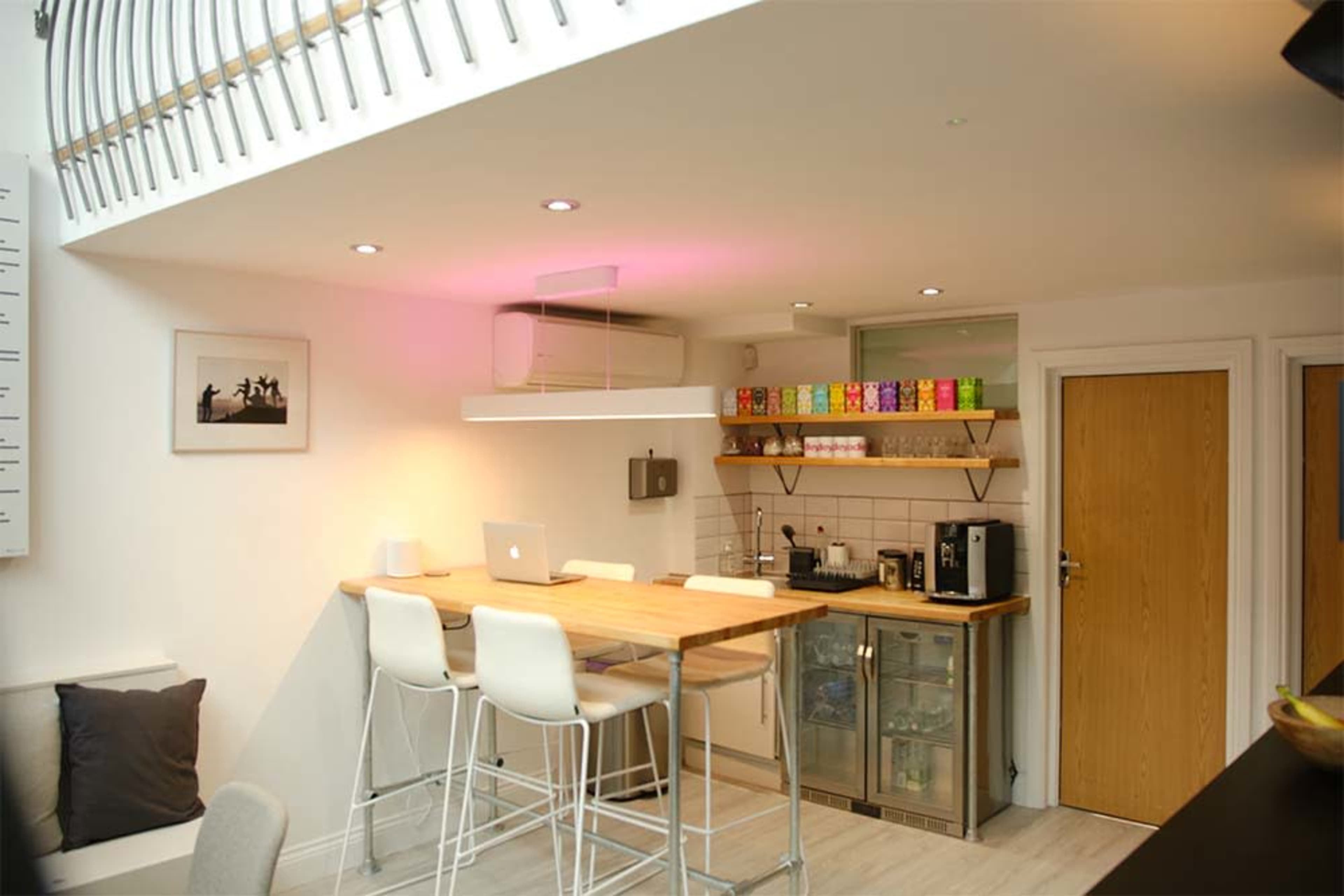 A modern kitchen with a wooden bar table, high stools, and a shelf displaying colorful boxes, illuminated by recessed lighting.