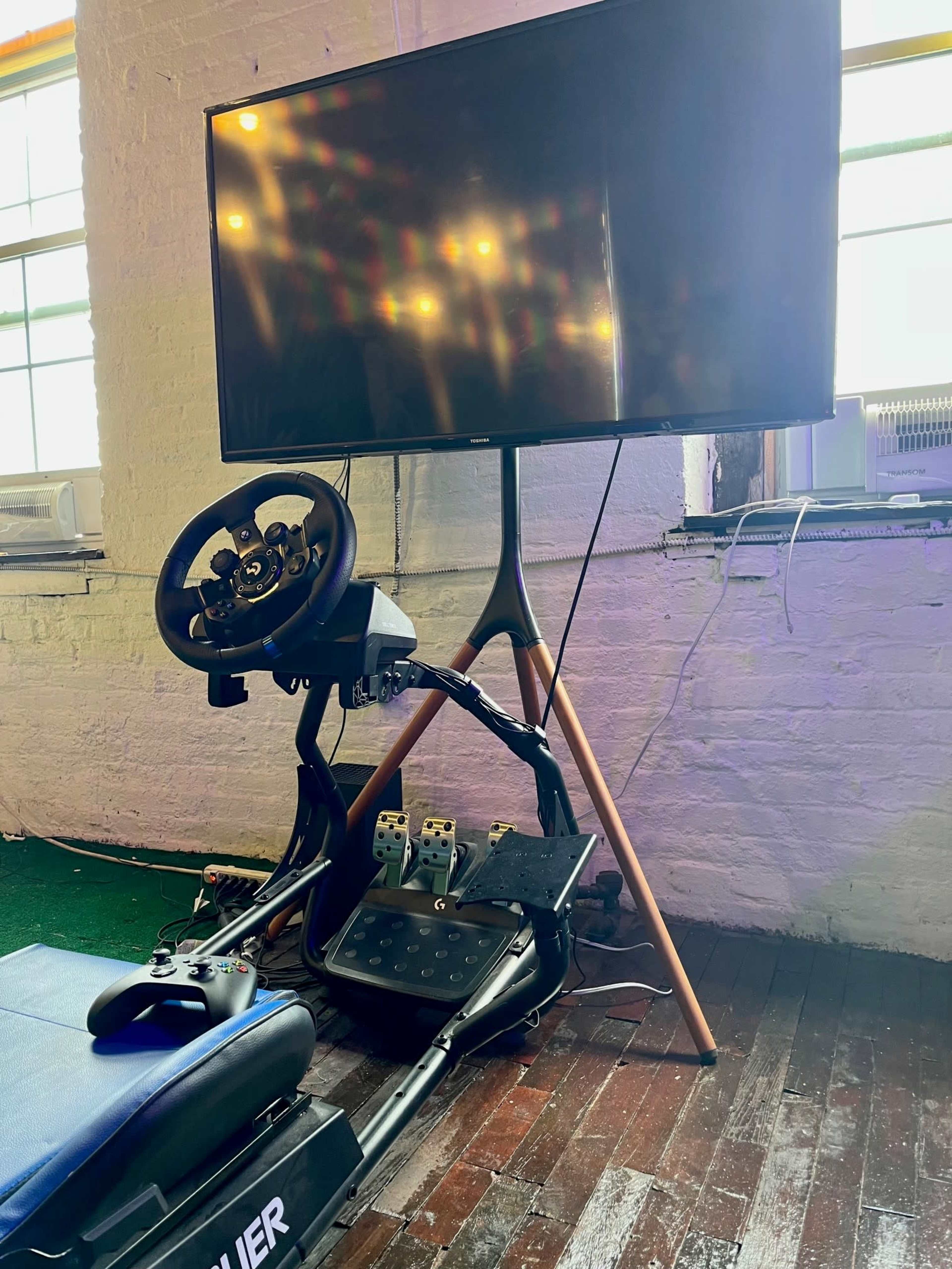 A racing simulation setup features a steering wheel and pedal system alongside an Xbox controller, positioned near a large flat-screen TV in a room with wooden flooring.