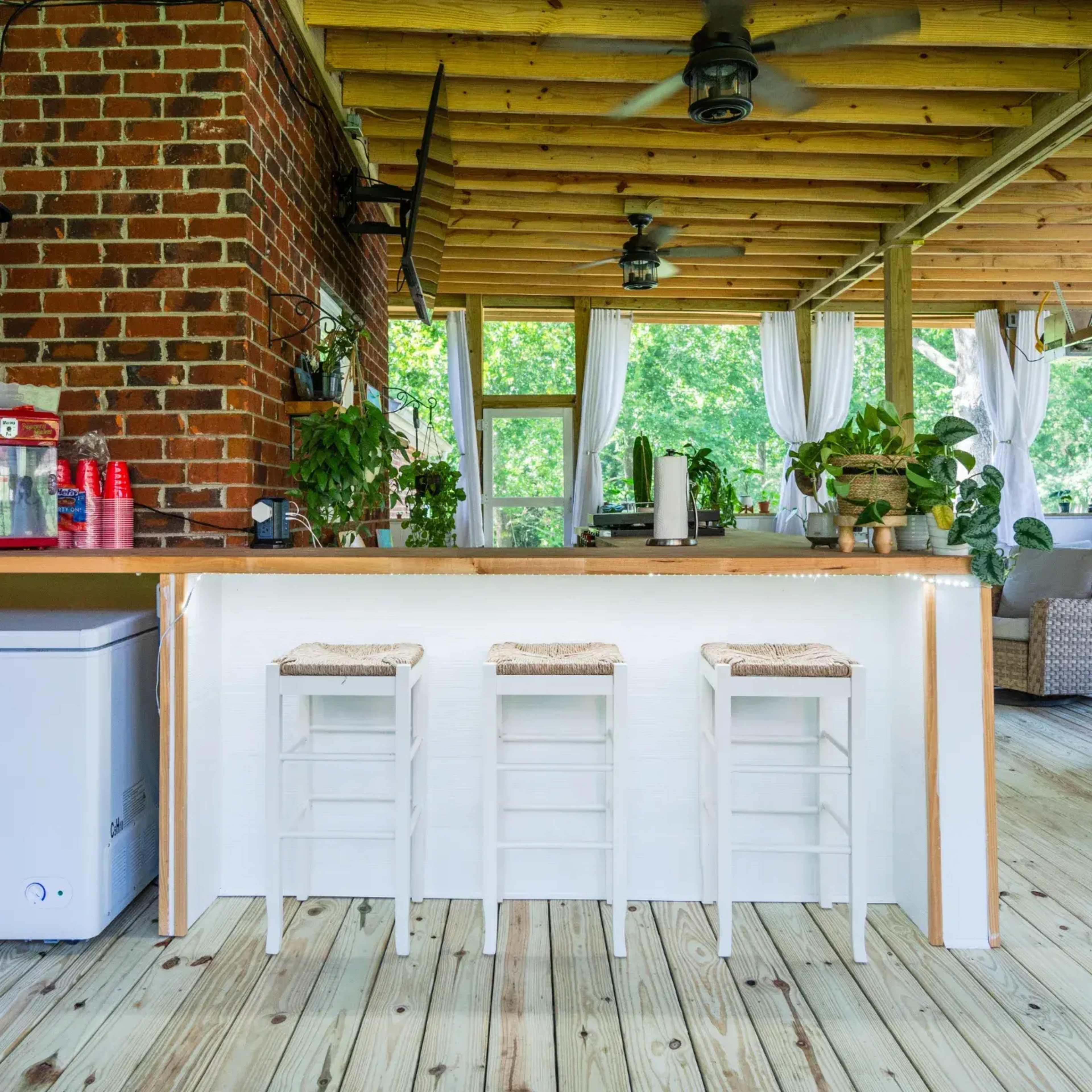 The image shows a rustic outdoor kitchen area featuring a wooden bar with three white stools, brick walls, and lush greenery in the background.