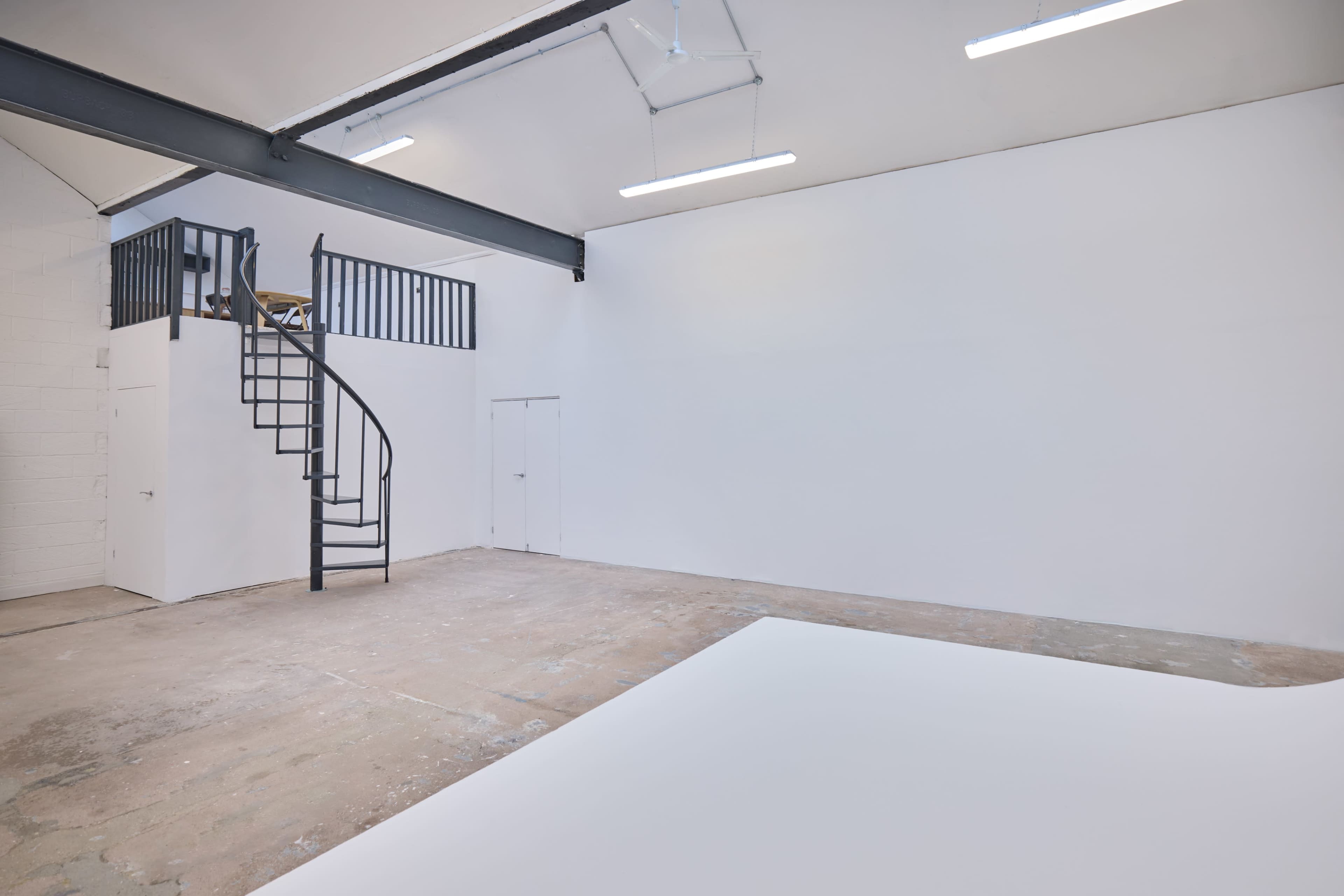The image shows an empty interior space with a spiral staircase leading to a mezzanine, characterized by white walls and a concrete floor.