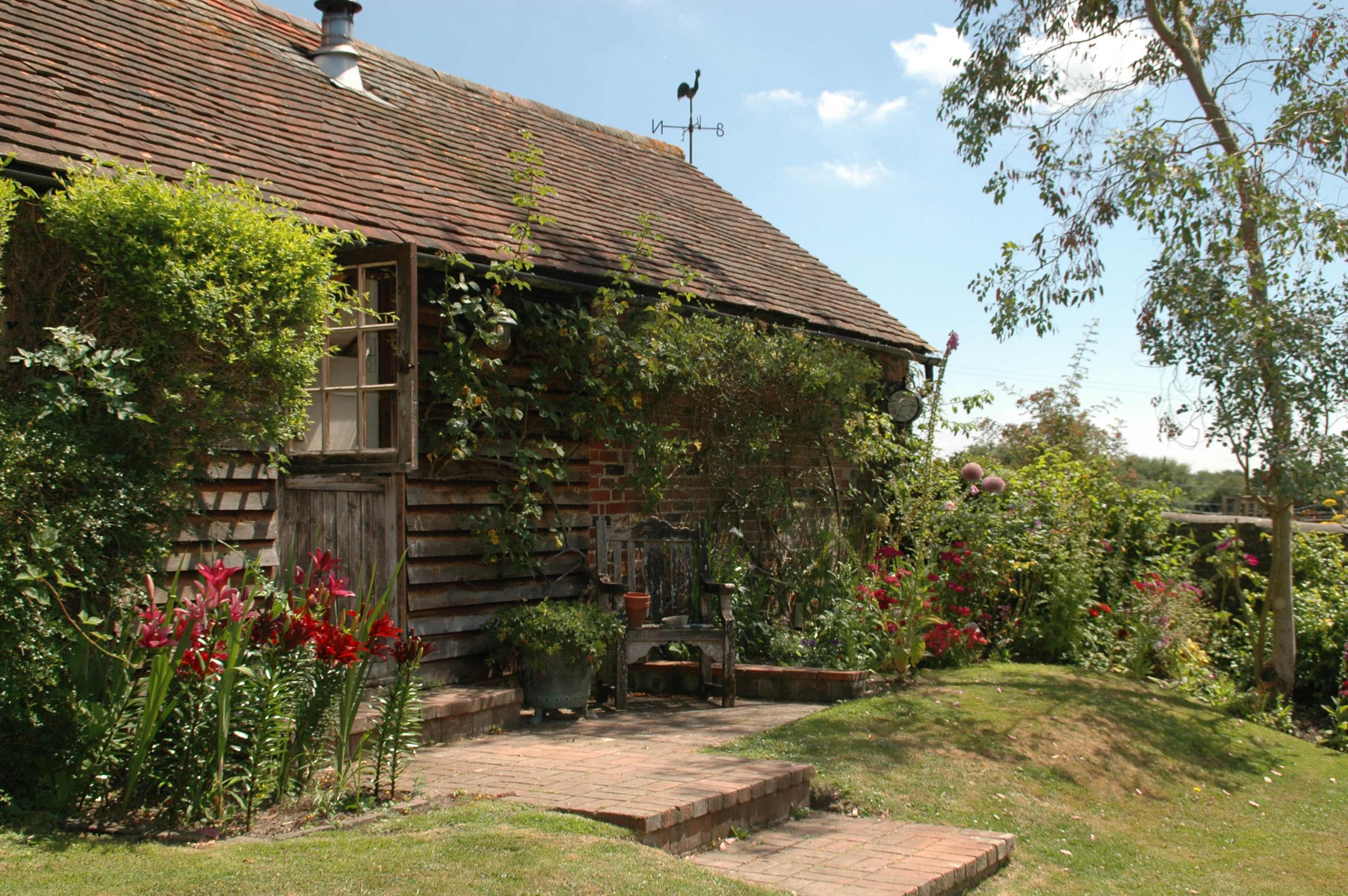 A rustic wooden cottage, surrounded by vibrant flowers and greenery, features a welcoming entrance with stone steps.