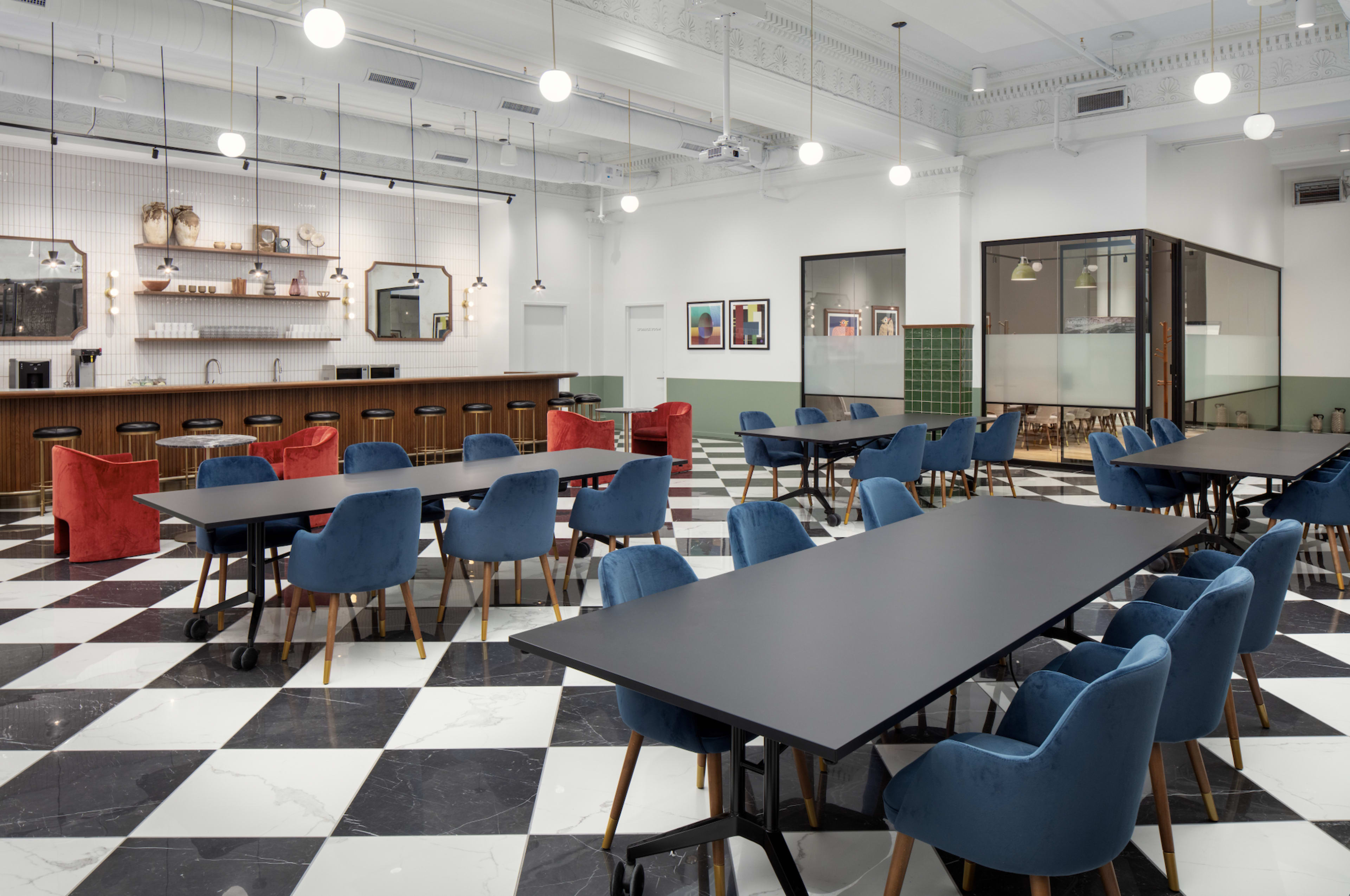 A modern, spacious dining area with checkered black and white flooring, featuring several tables surrounded by blue and red chairs, and a bar counter in the background.