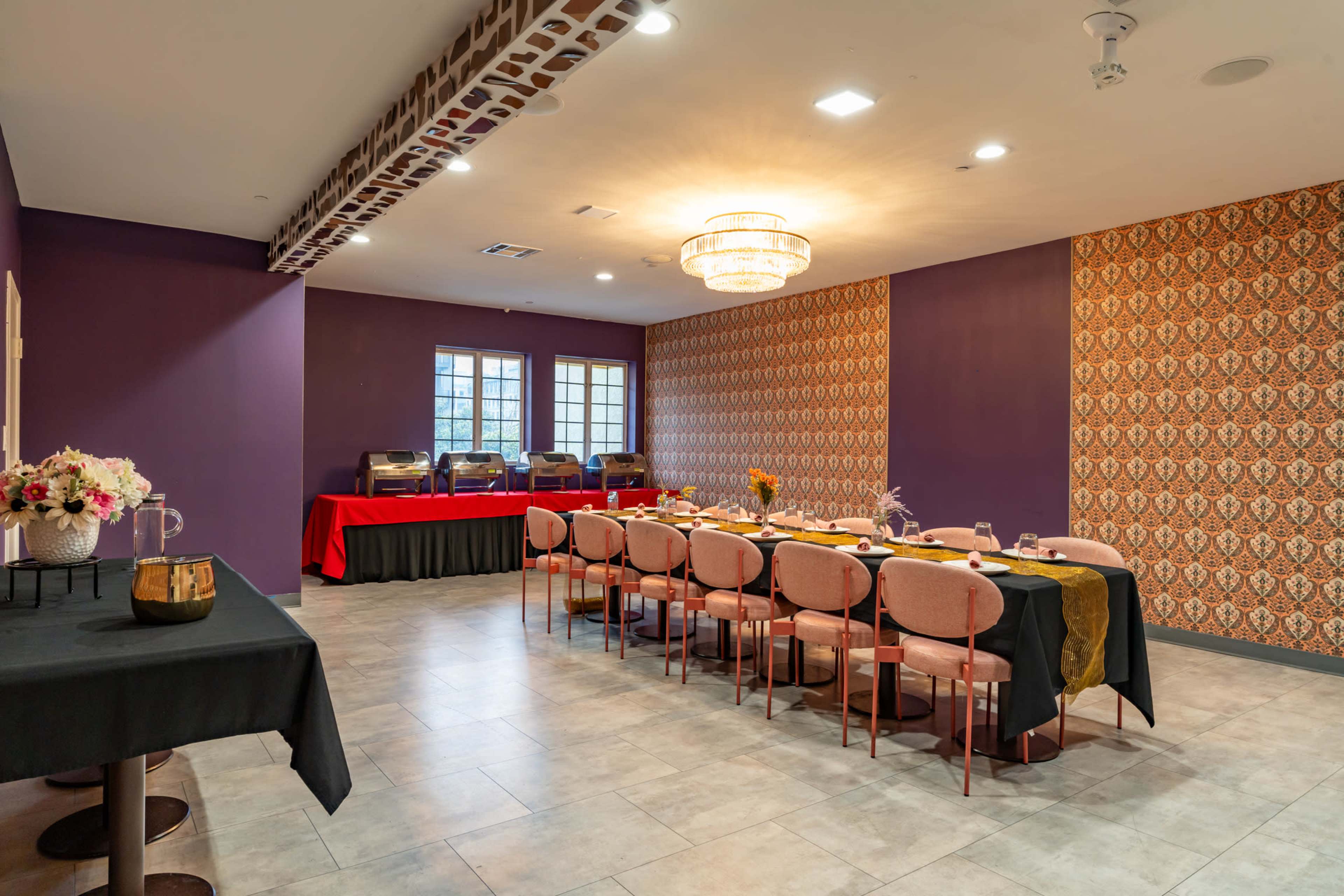 The image shows a dining room with a long table set for a meal, surrounded by pink chairs, a dark table in the foreground, and decorative wallpaper on one wall.