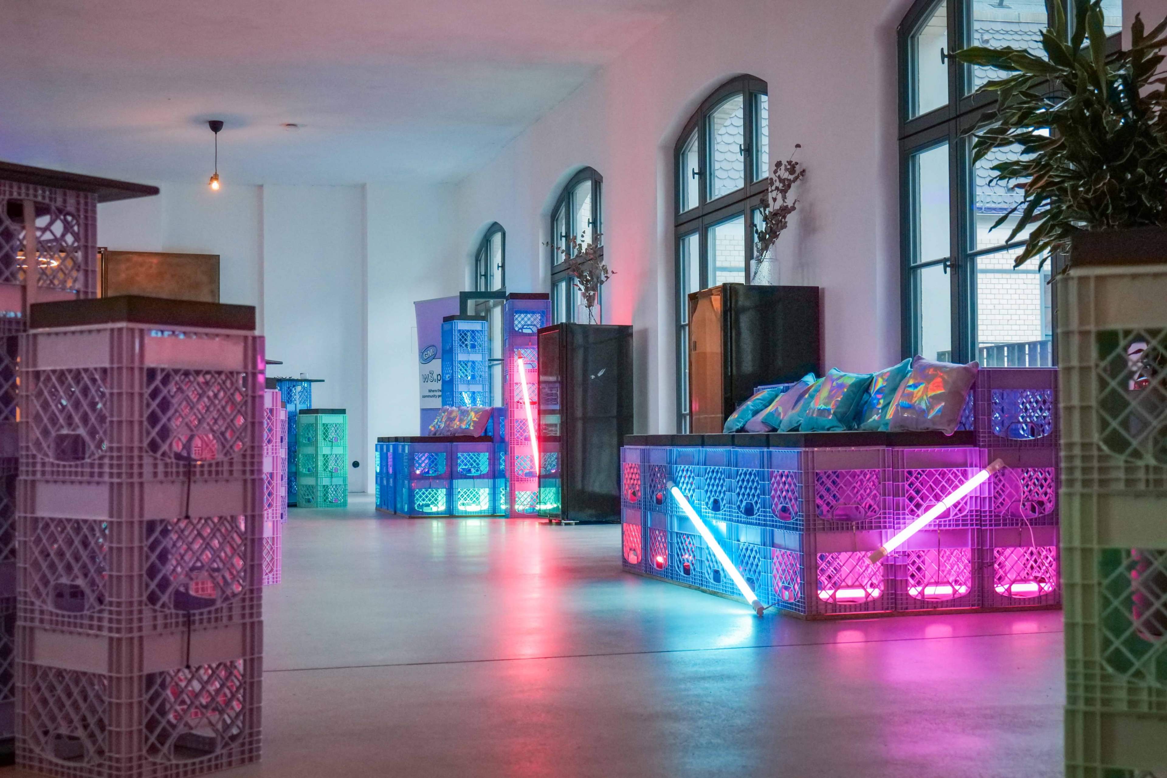 The image shows a room arranged with colorful crates serving as furniture, illuminated by vibrant lighting.