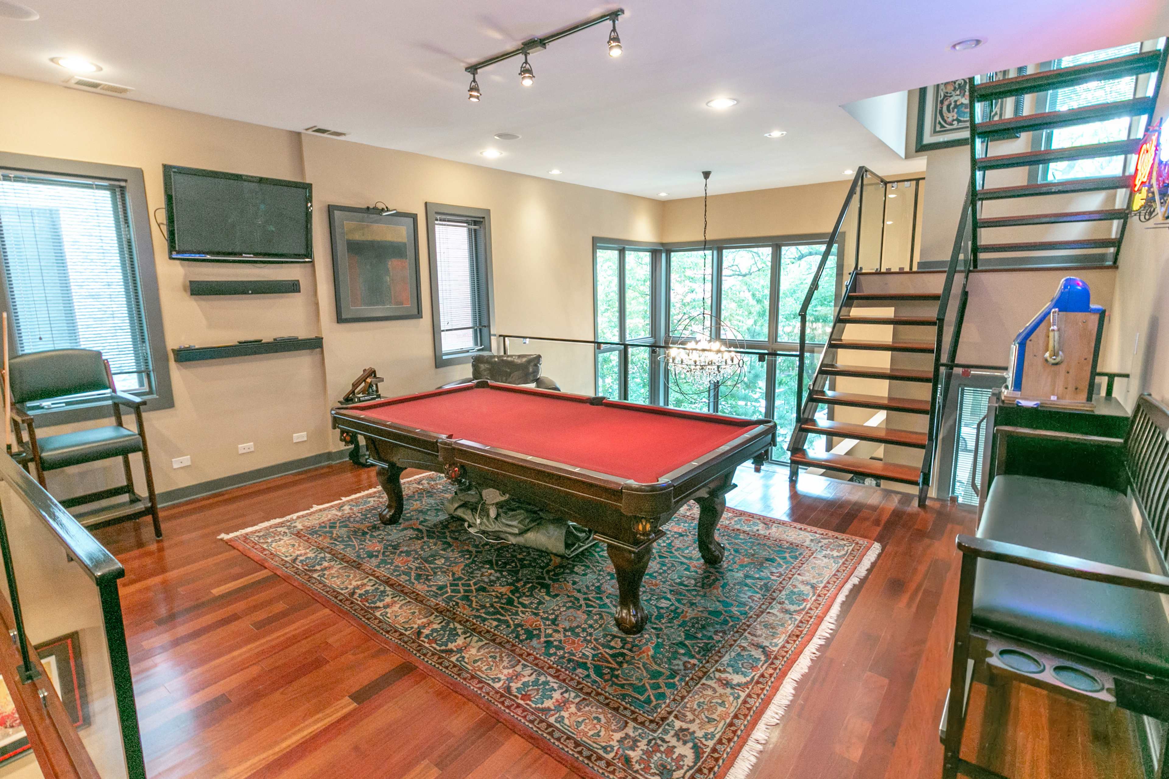 The image shows a modern game room with a red pool table, a staircase, large windows, and hardwood flooring.