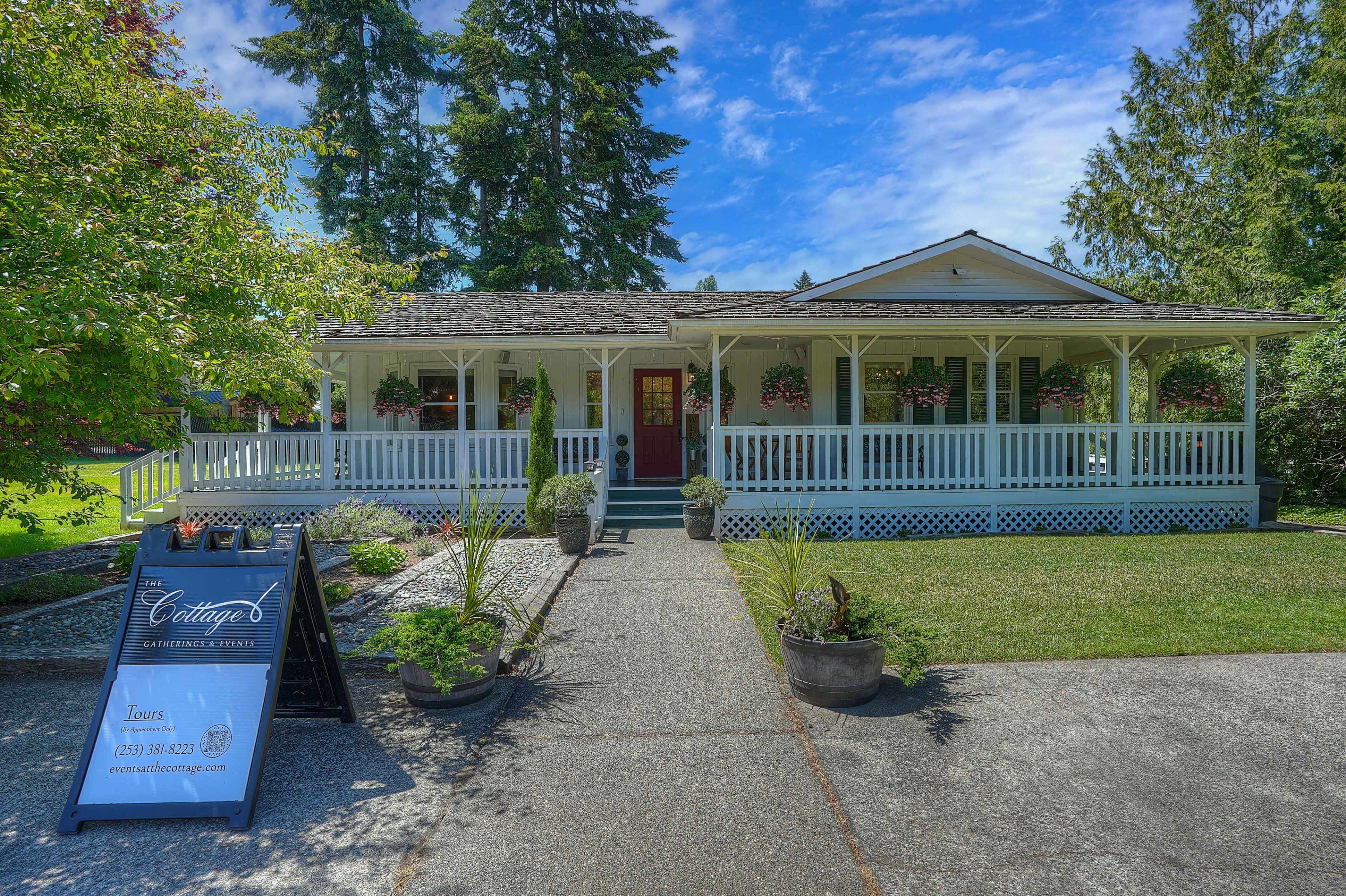 The image shows a single-story cottage surrounded by greenery, featuring a front porch with hanging flower baskets and a signboard nearby.