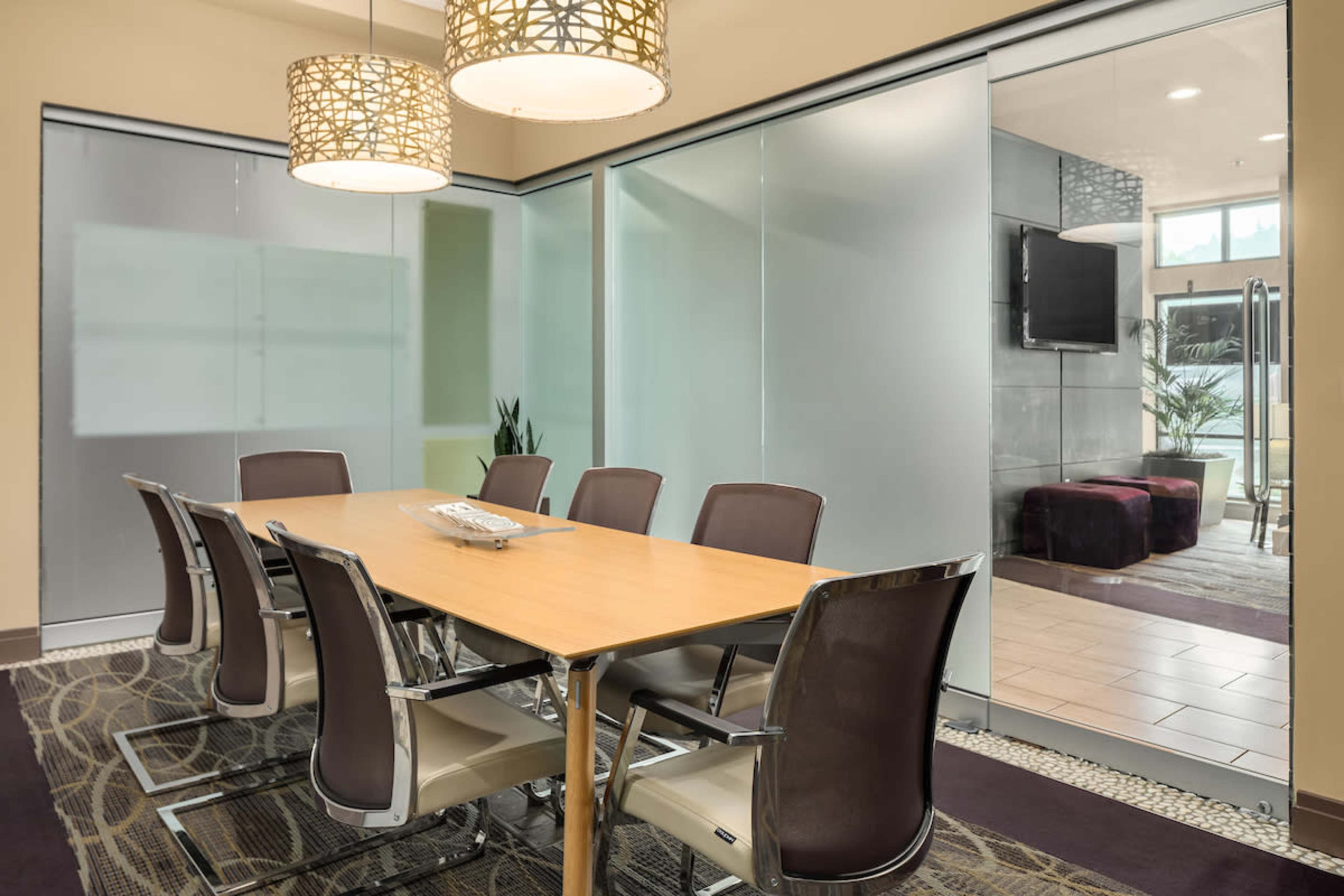 A modern conference room features a large wooden table surrounded by eight chairs, with glass walls and pendant lighting.