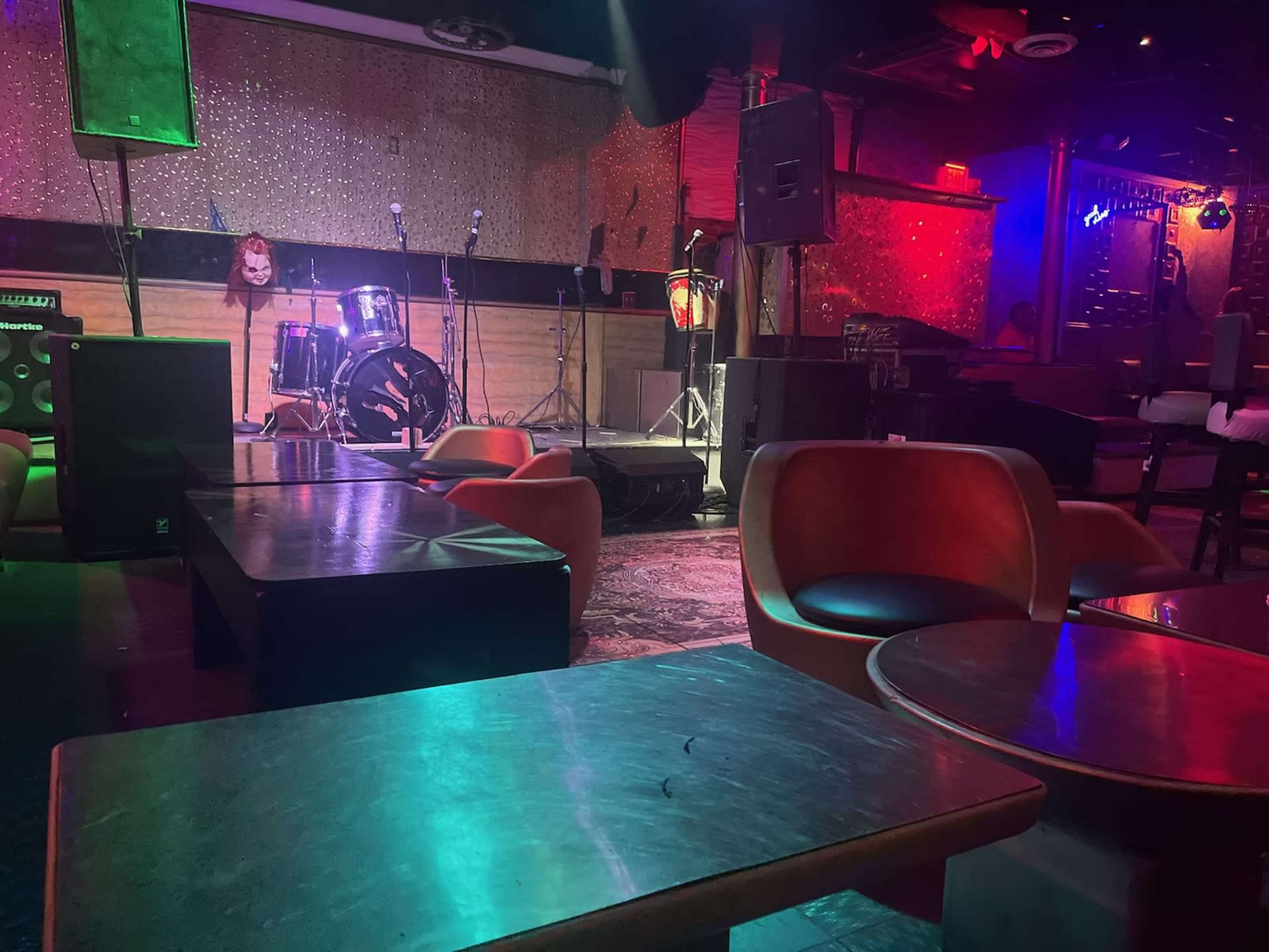 The image shows a dimly lit music venue with empty tables and a stage set up with a drum kit in the background.