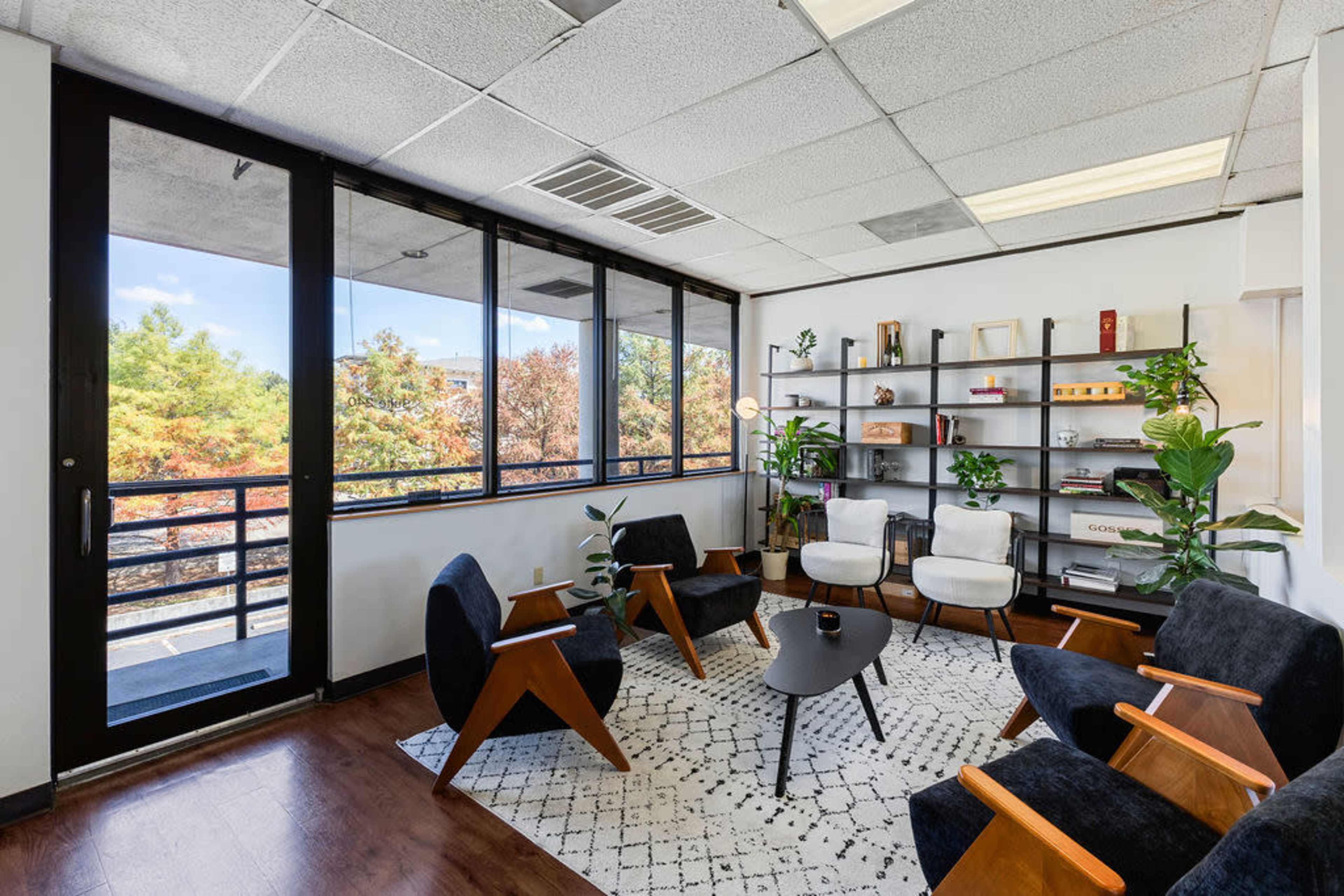 The image depicts a modern office waiting area featuring a black and white rug, four dark armchairs, a round coffee table, and a large window with a view of trees.