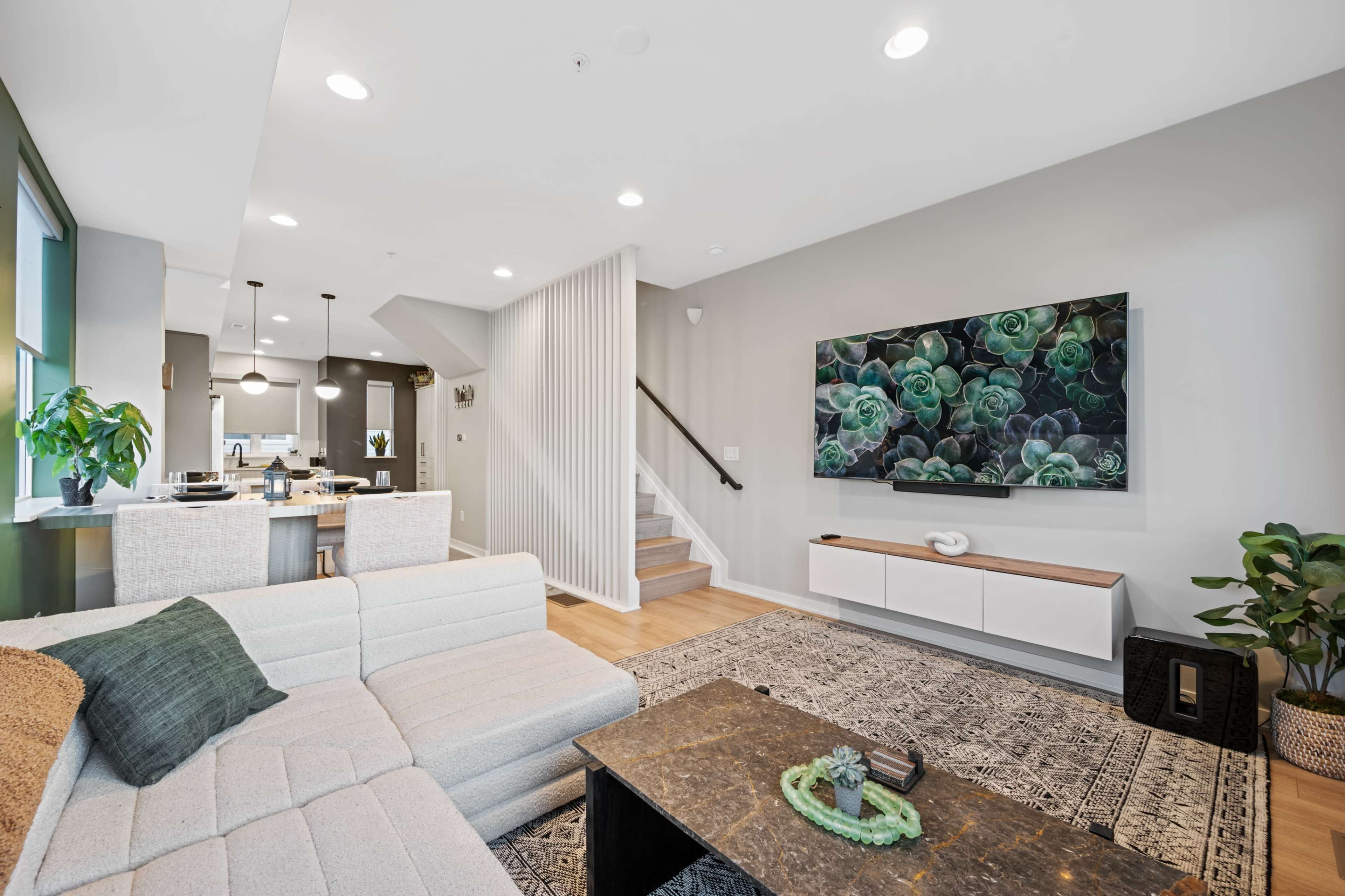 The image shows a modern living room featuring a sectional sofa, a coffee table, a wall-mounted television displaying a botanical image, and a dining area visible in the background.