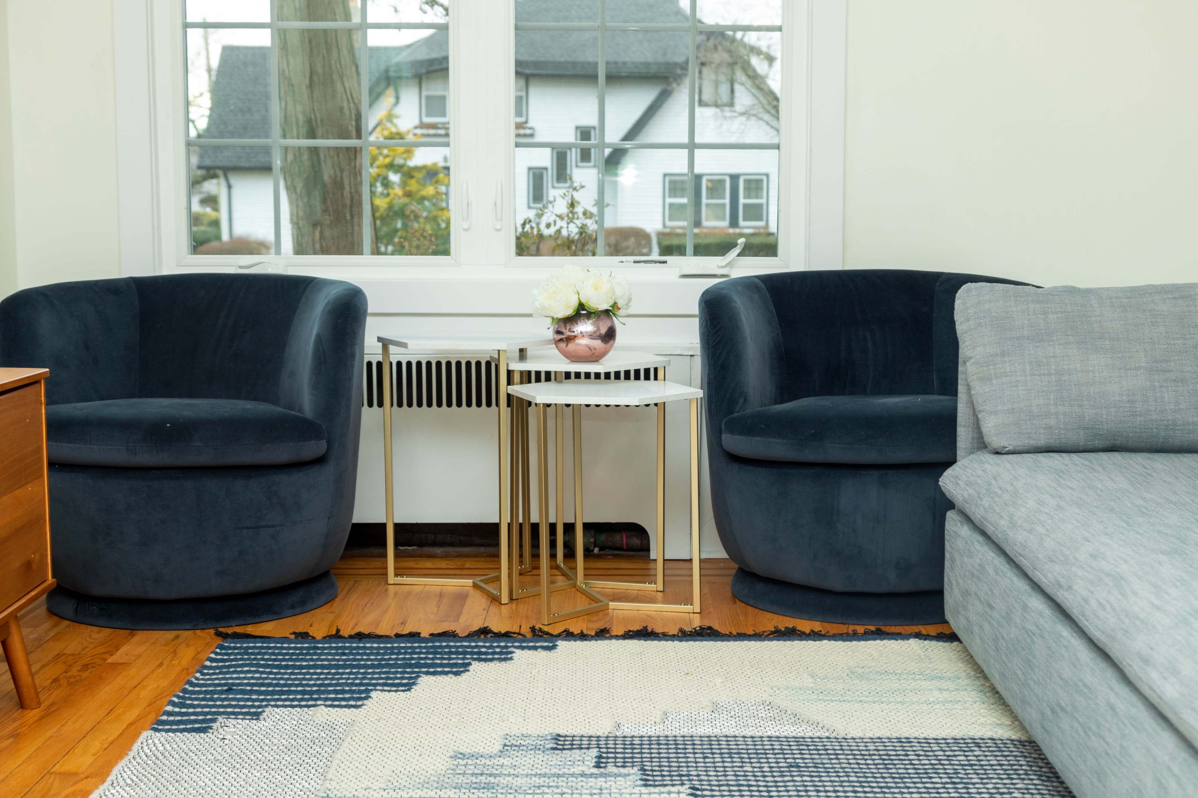 The image shows a living room with two velvet chairs, a small white and gold side table, and a gray sofa, all set near a large window overlooking a house and trees outside.