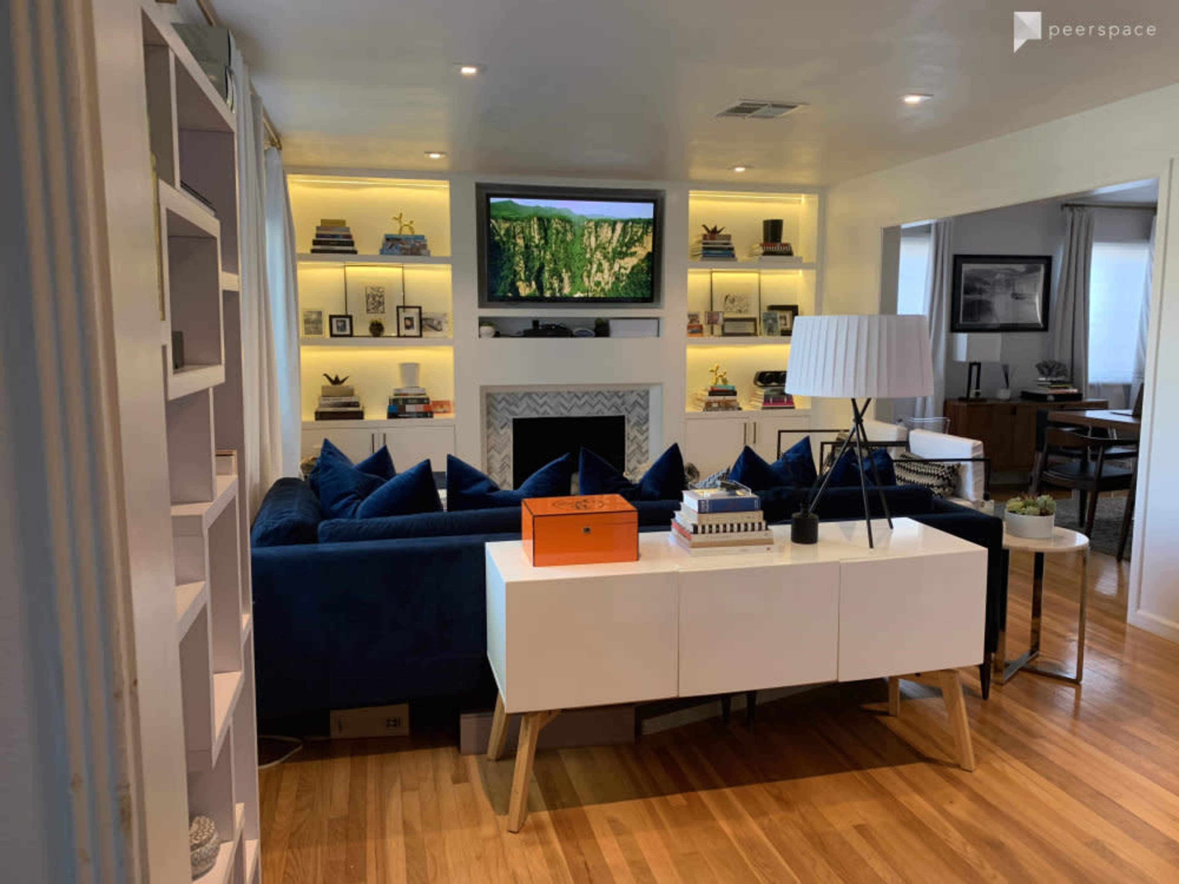A living room with a blue sectional sofa, a white console table, a mounted television above a fireplace, and built-in shelves displaying books and decor.