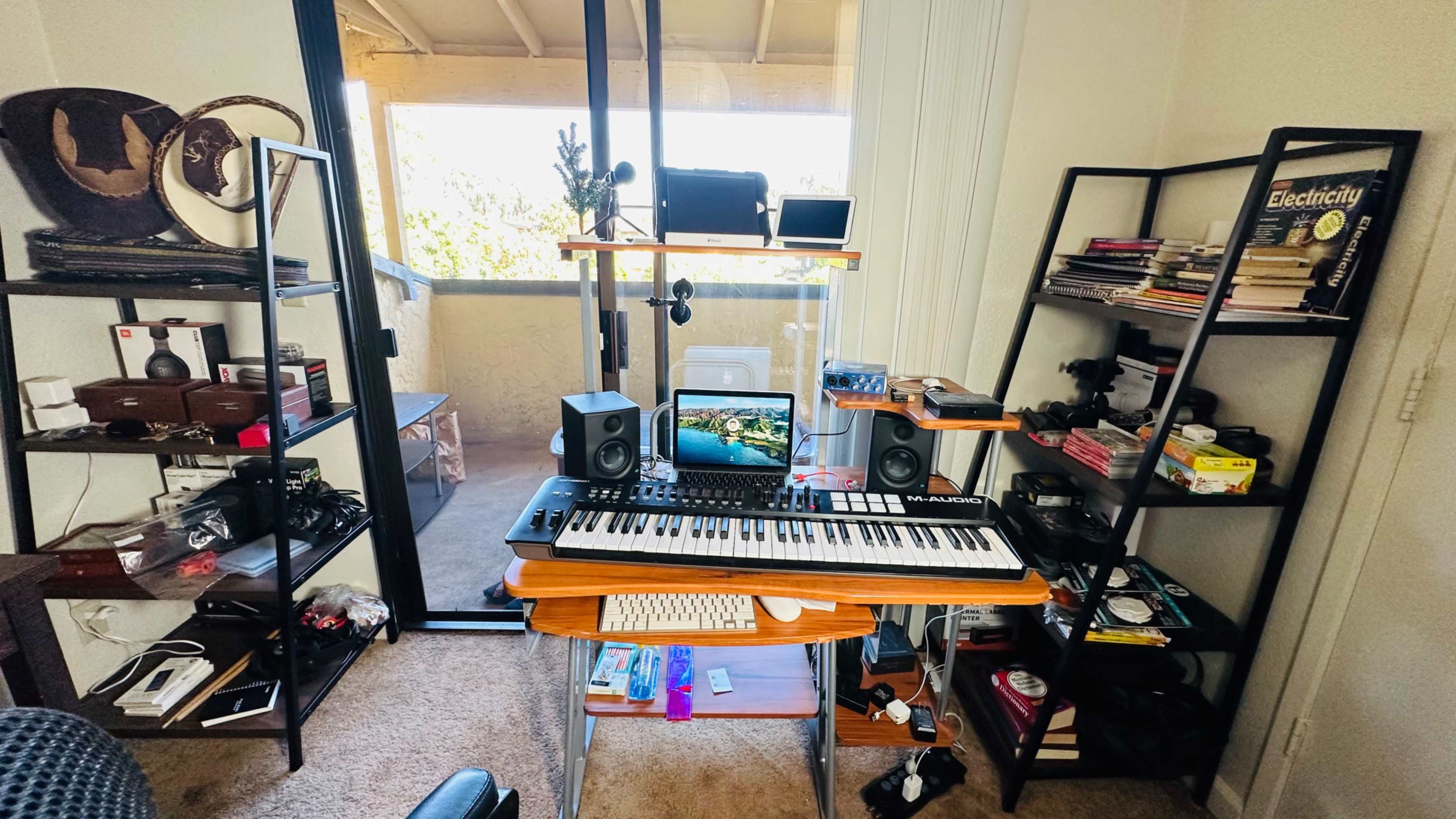 A home office setup features a keyboard on a desk with speakers and a computer monitor, surrounded by shelves filled with books and various equipment.