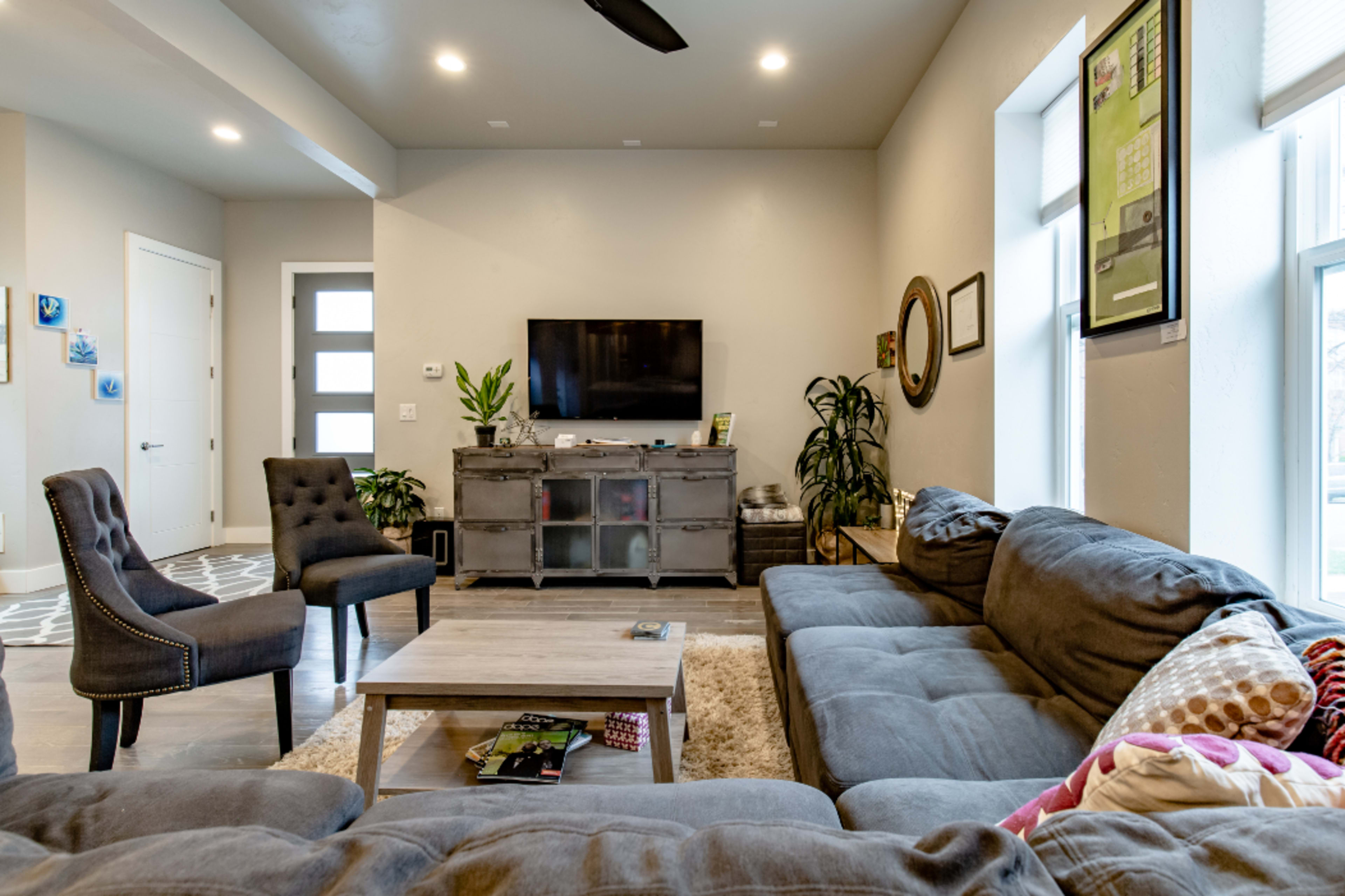 The living room features a large gray sectional sofa, a wooden coffee table, two stylish chairs, and a television mounted on the wall, surrounded by plants and decor.