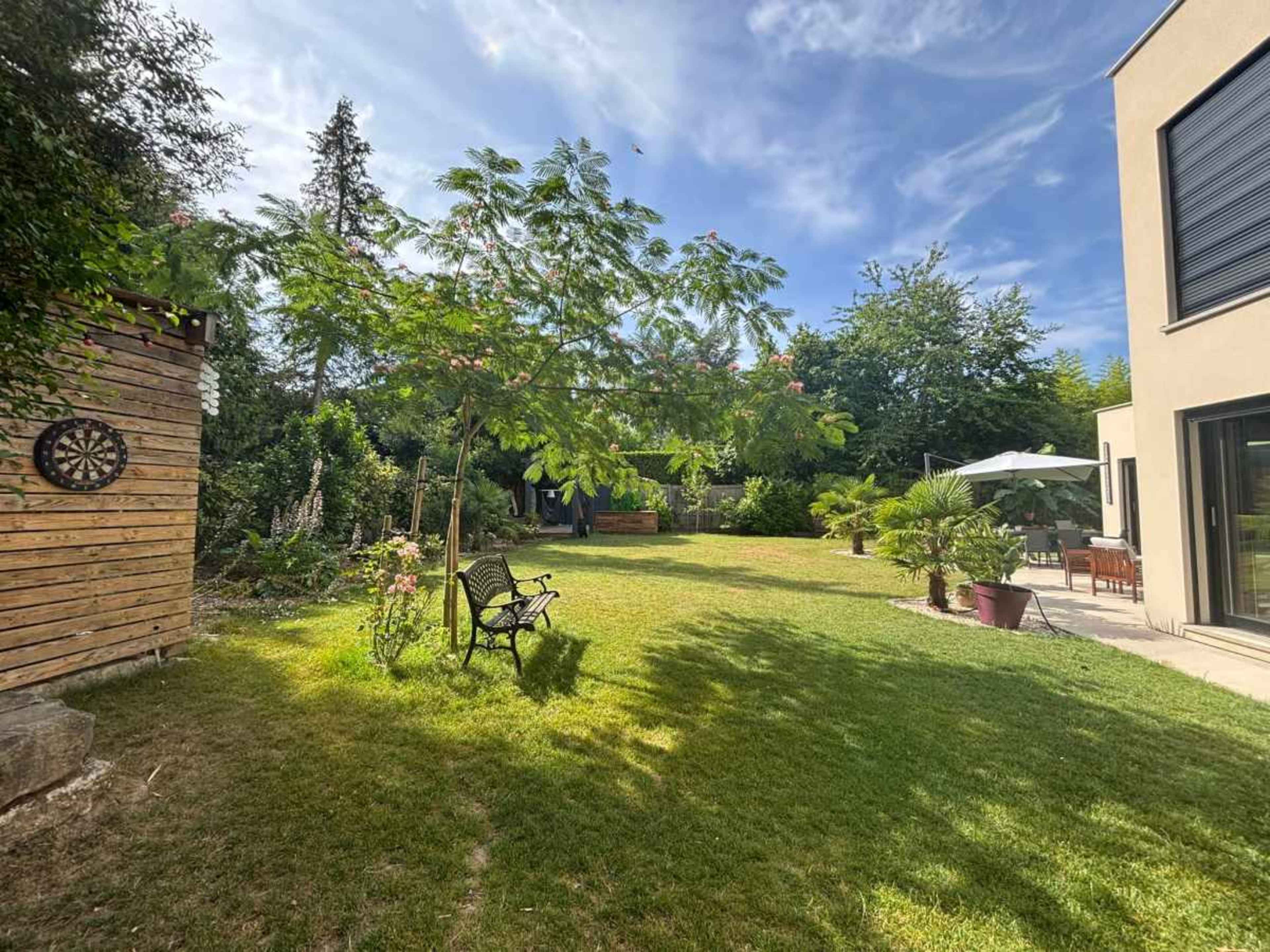 The image shows a spacious garden with a grassy area, a wooden structure on the left, and patio furniture near a modern house on the right.