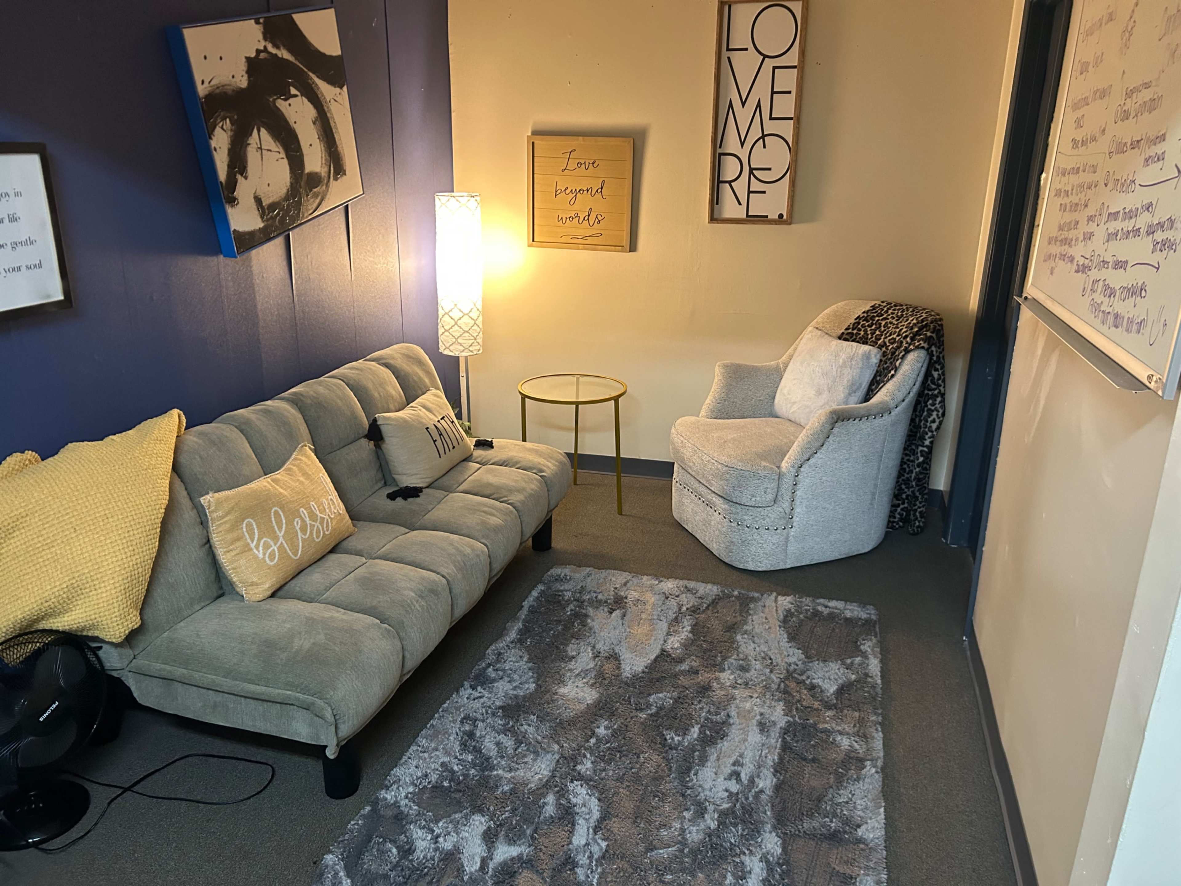 The image shows a cozy room with a gray couch, a light-colored armchair, a small round table, and wall art, illuminated by a floor lamp.