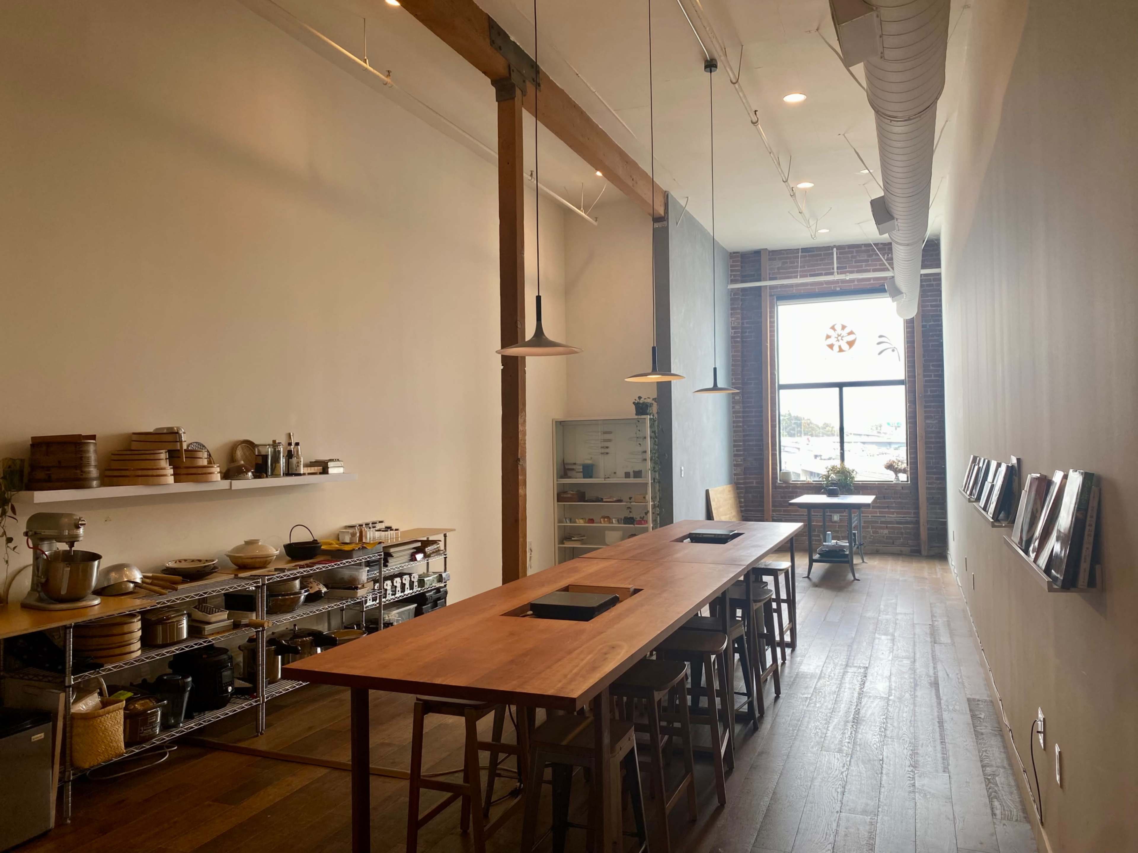 The image shows a spacious kitchen with a long wooden table, metal shelves filled with kitchenware, and a large window providing natural light.