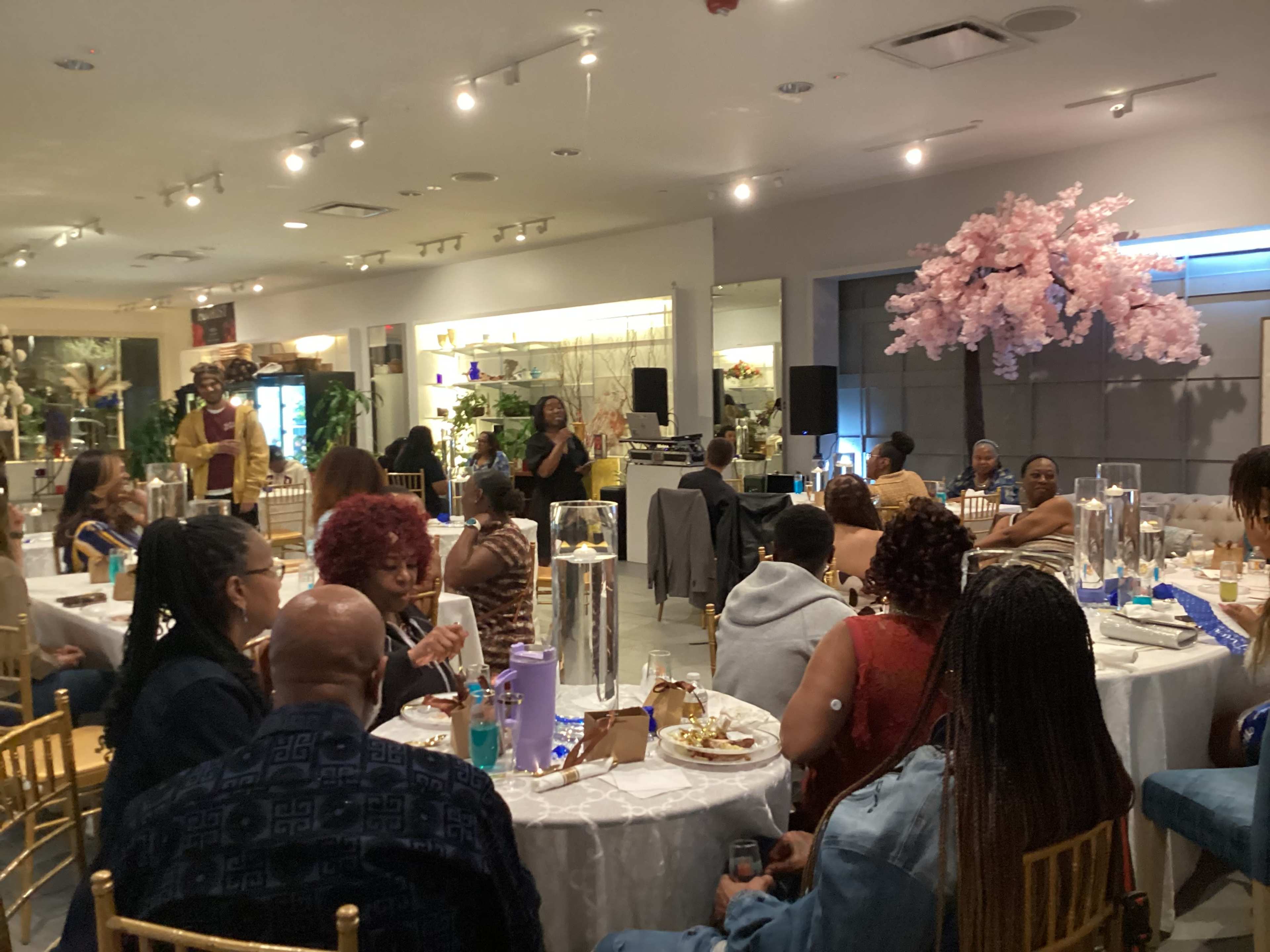 The image shows a dining event with guests seated at round tables adorned with white tablecloths and floral centerpieces, while a speaker addresses the audience in a well-lit space.