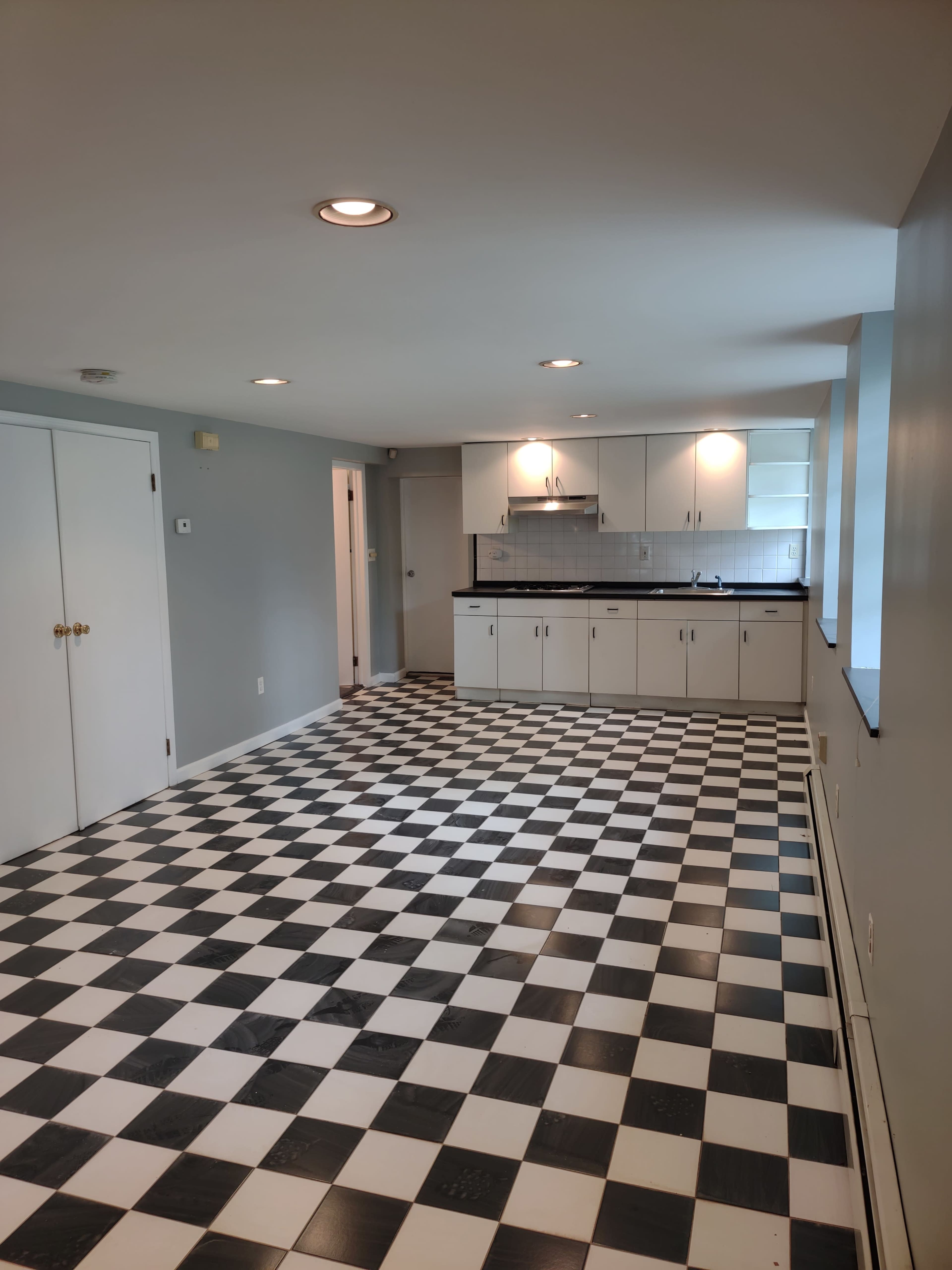 A spacious, open room featuring a black and white checkered floor and a kitchen area with white cabinets and a black countertop.