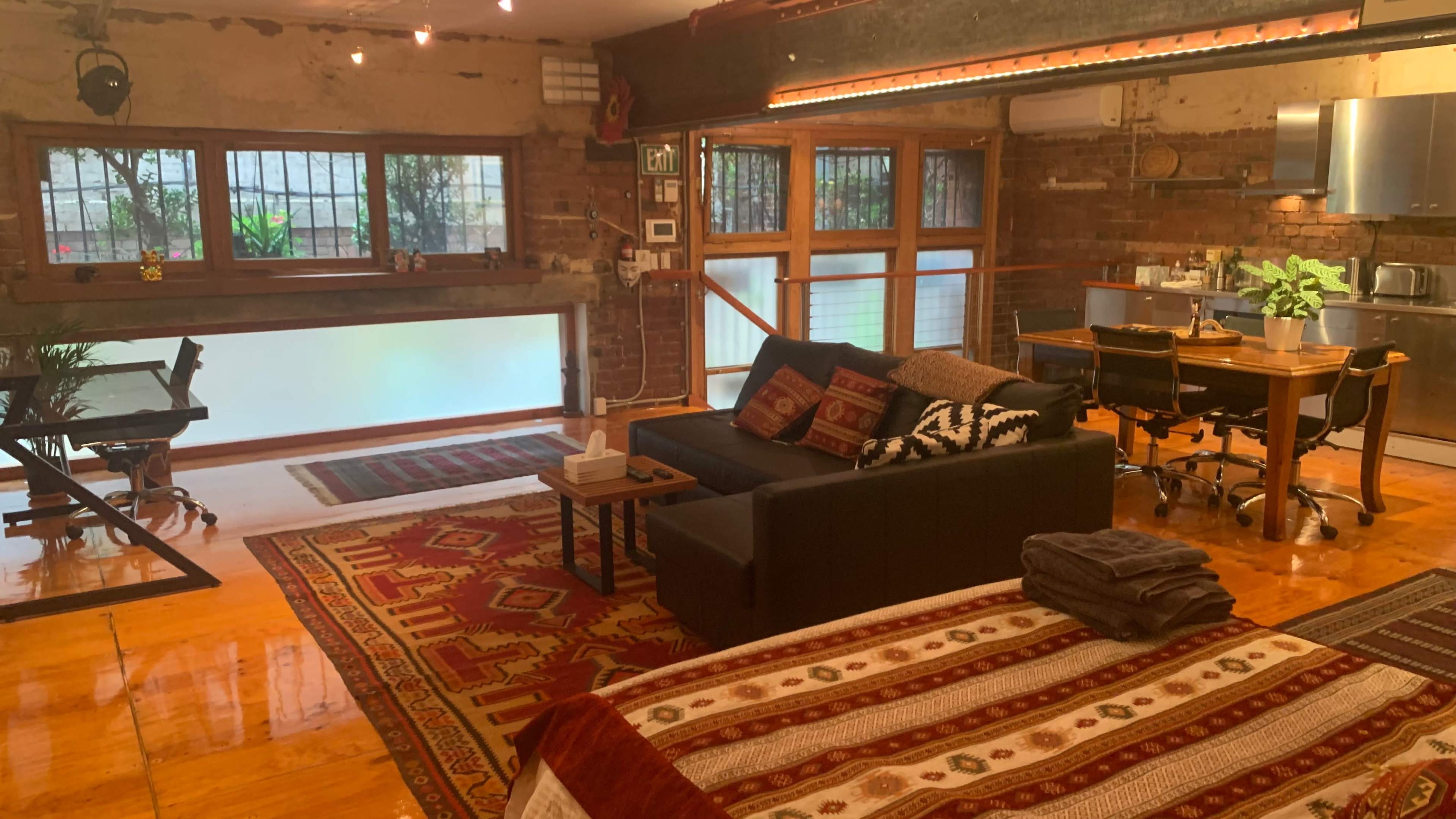 A stylish loft with exposed brick walls, hardwood floors, and a living area featuring a sofa, a dining table, and a workspace beside large windows.