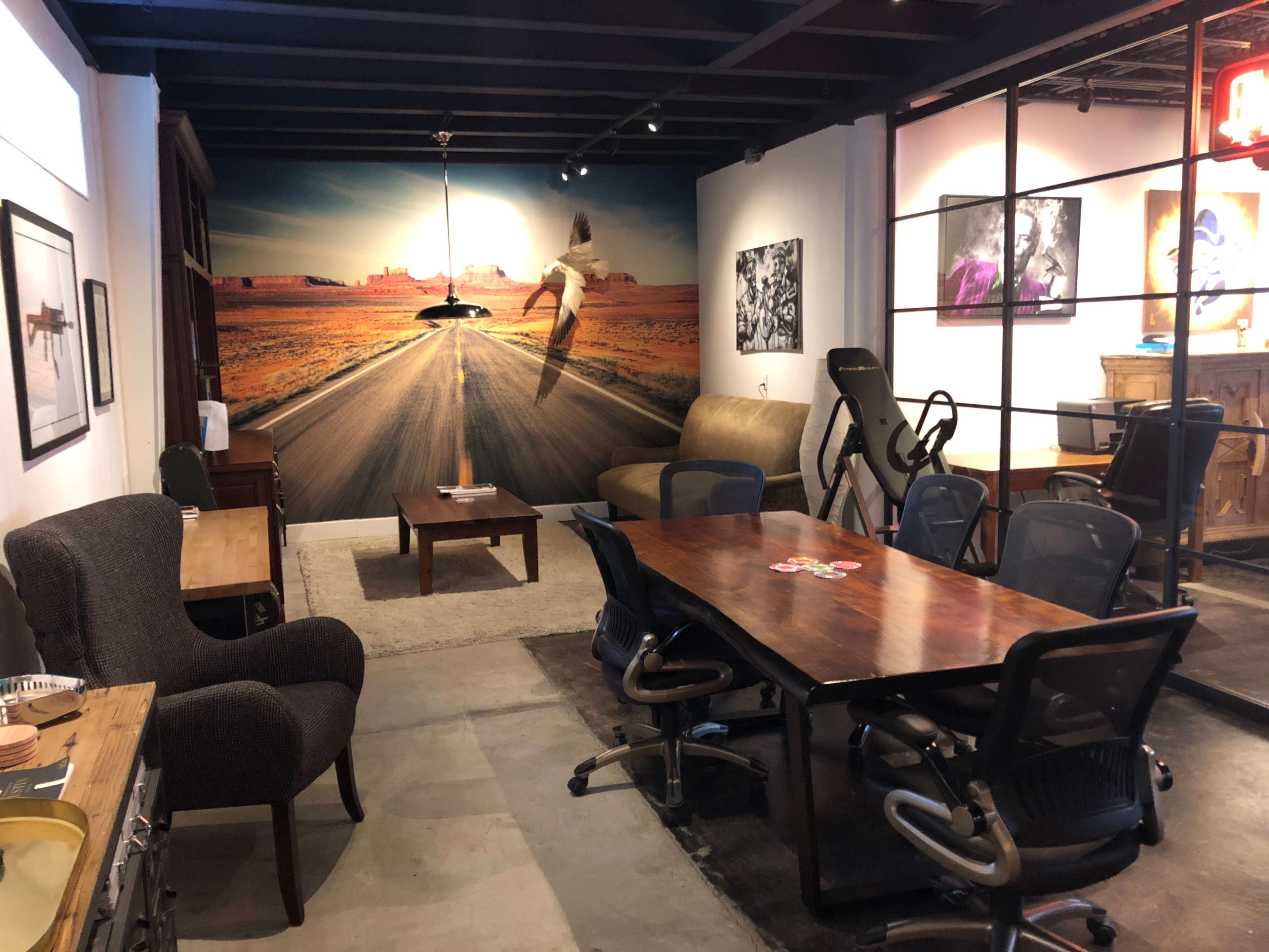 The image shows a modern office space featuring a large wall mural of a desert landscape, a wooden dining table surrounded by office chairs, and a seating area with plush chairs.