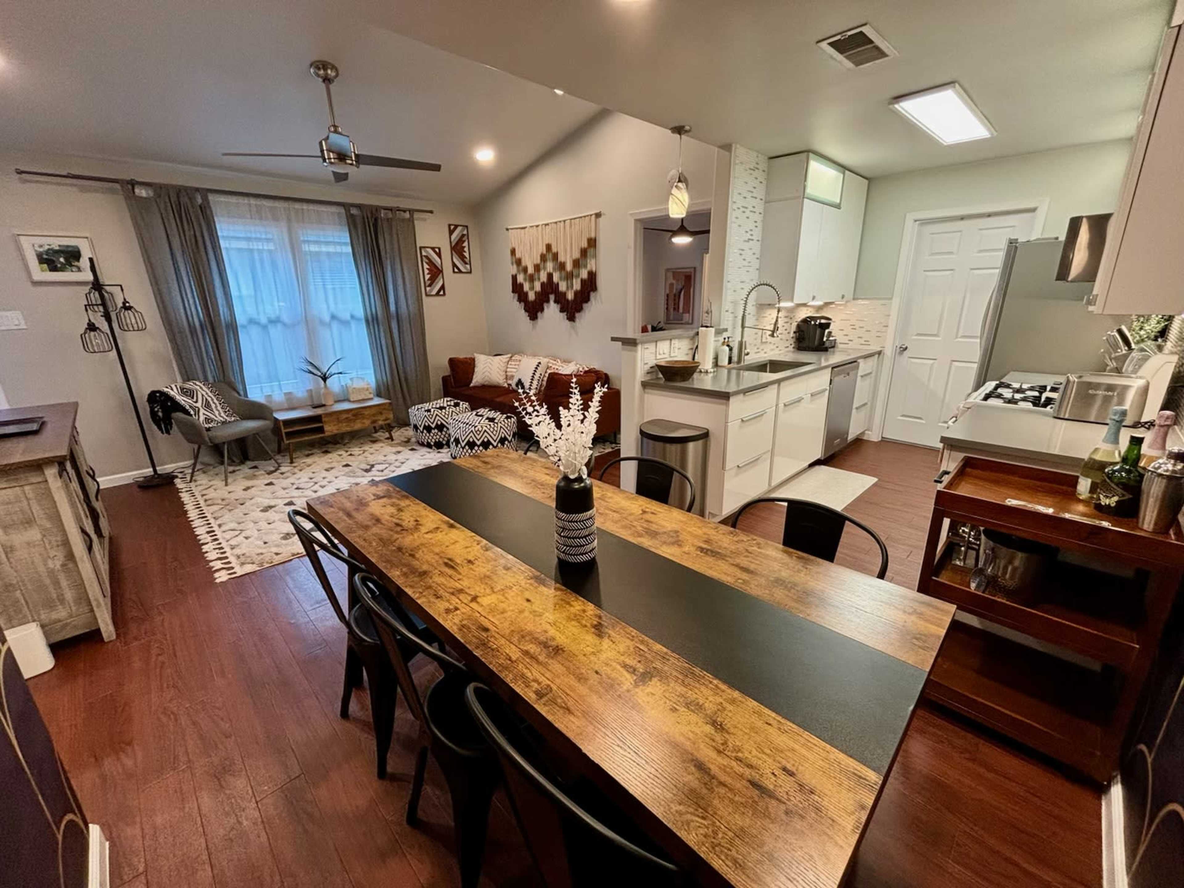 The image shows a modern open-concept kitchen and dining area with a wooden dining table, black chairs, and a living room featuring a couch and decorative accents.