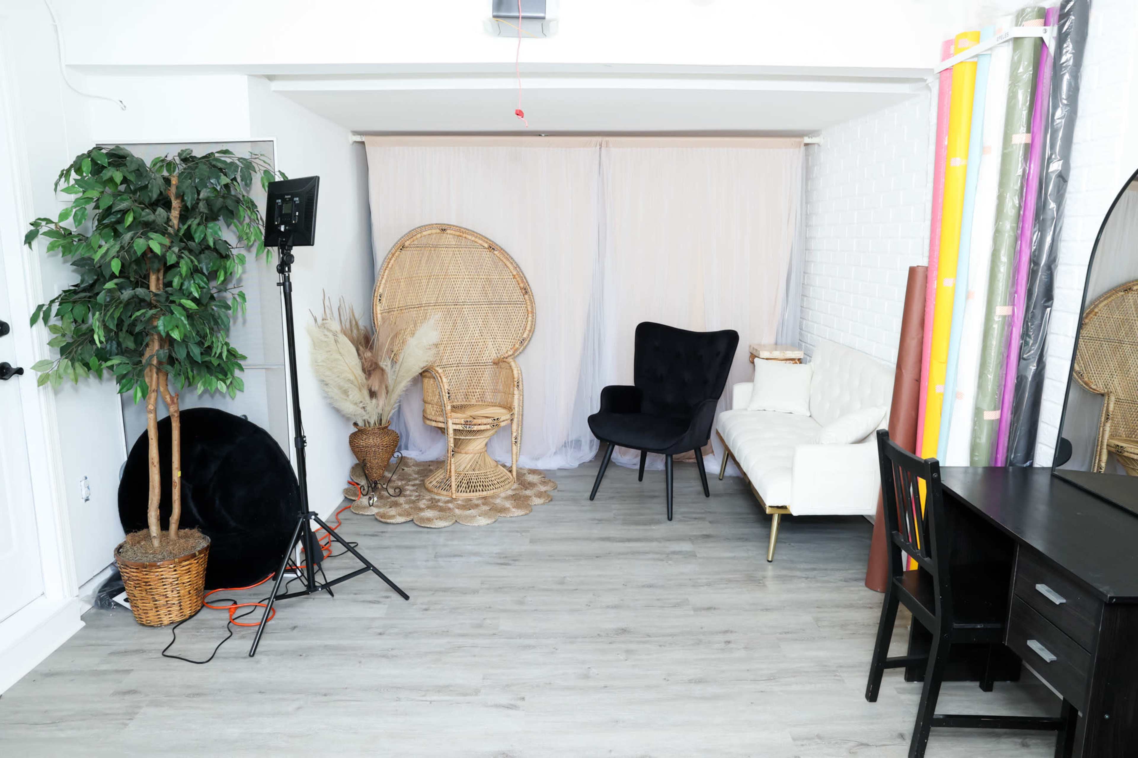 A styled interior space featuring a rattan chair, a black armchair, a white couch, potted plants, and colorful backdrops arranged along the walls.