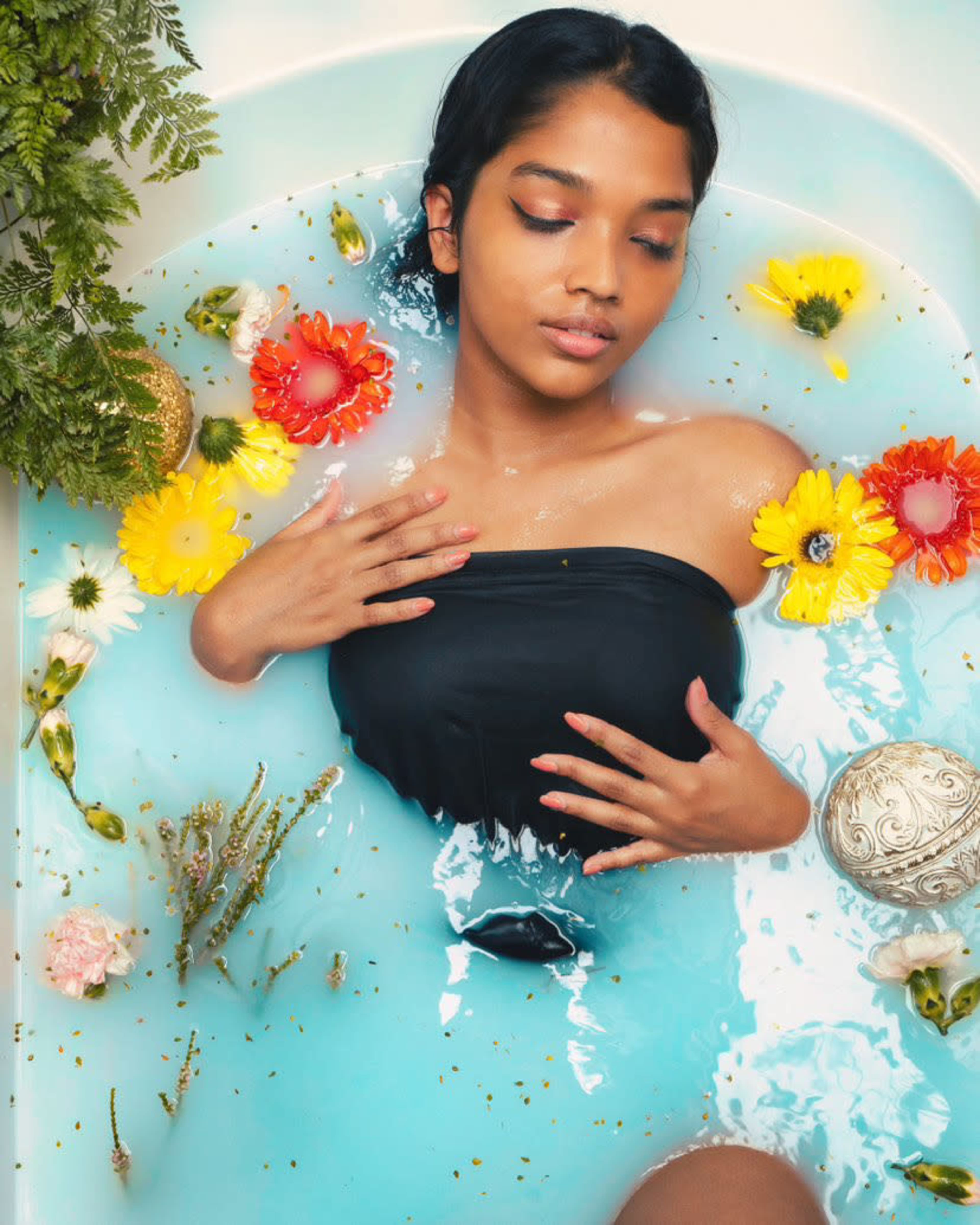 A person is relaxing in a bathtub filled with blue water, surrounded by colorful flowers and decorative elements.