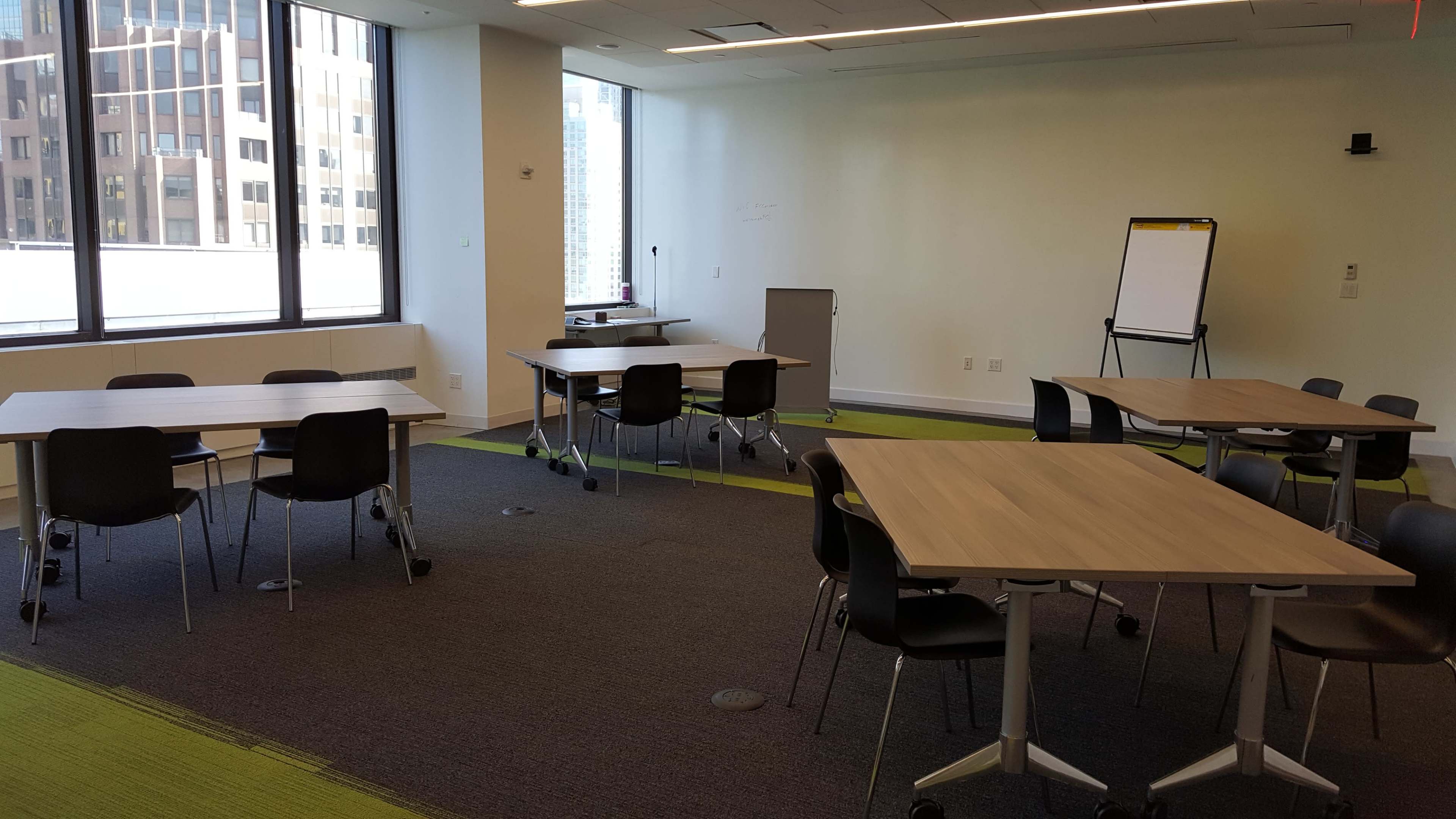 A conference room features multiple tables and chairs arranged on a carpeted floor, with large windows providing a view of a cityscape.