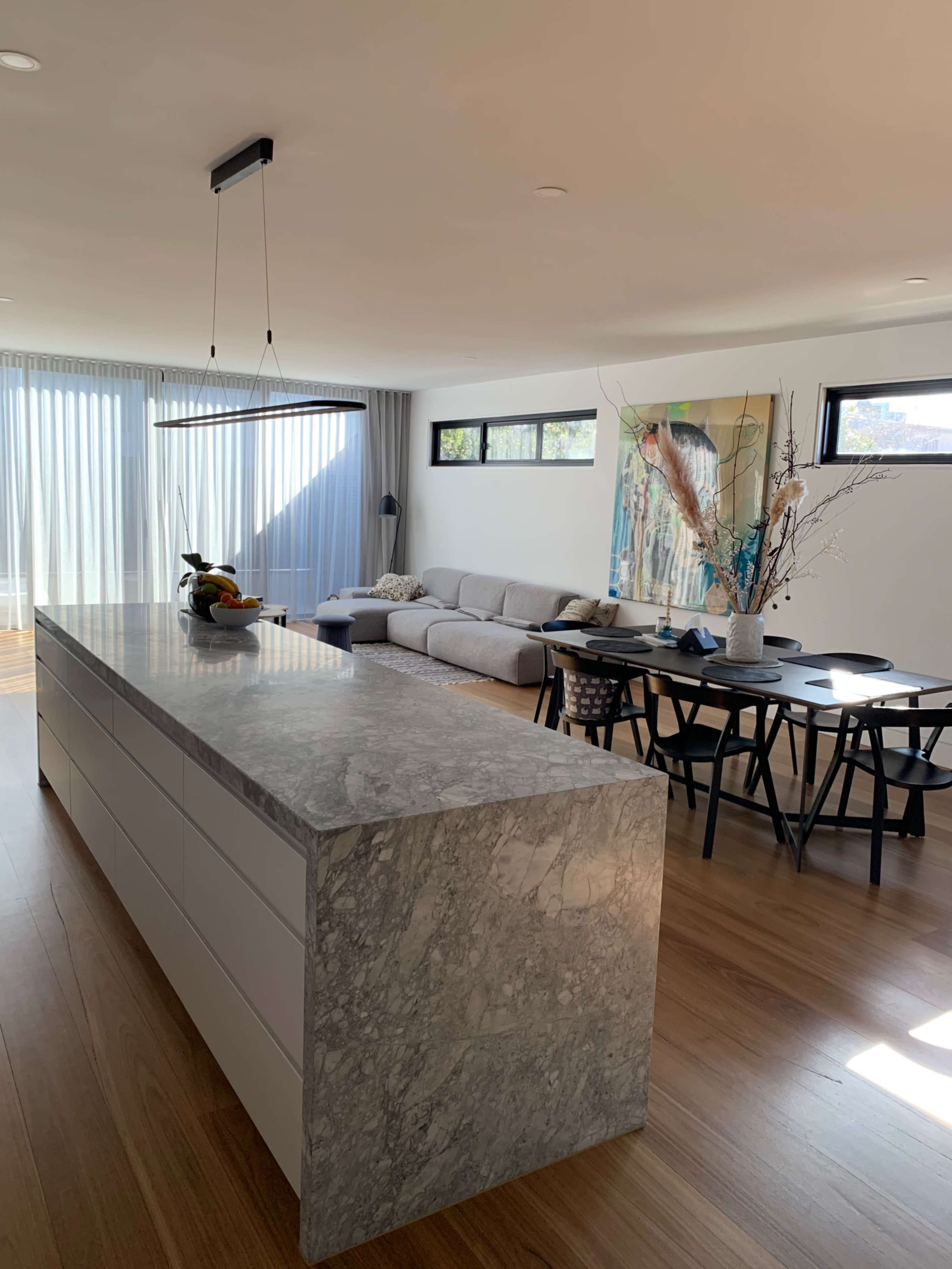 A modern open-concept kitchen and living area features a stone island, a grey sofa, a dining table, and large windows with sheer curtains.