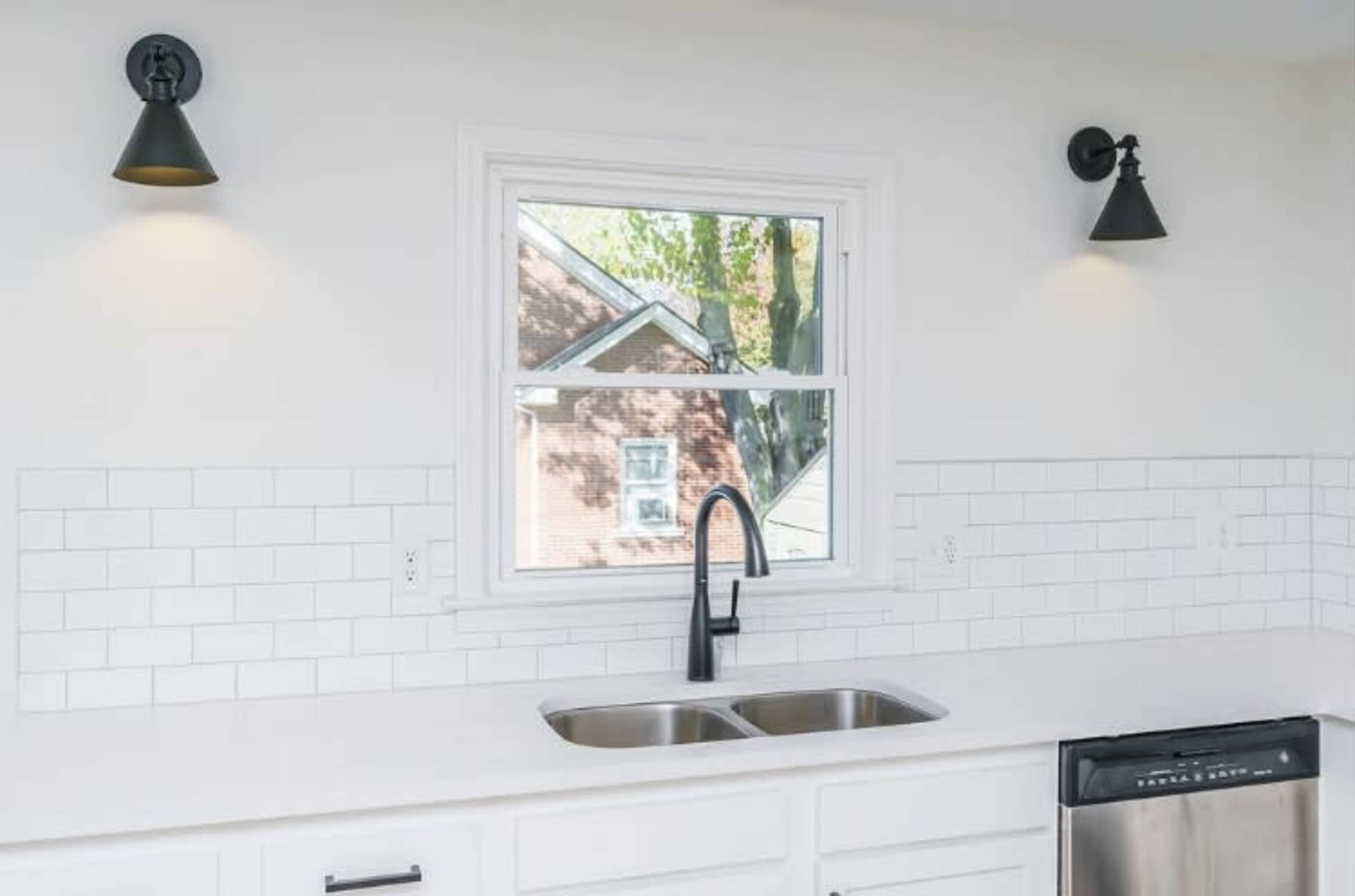 A modern kitchen with a white tiled backsplash, a double sink, and a window framed by wall sconces.