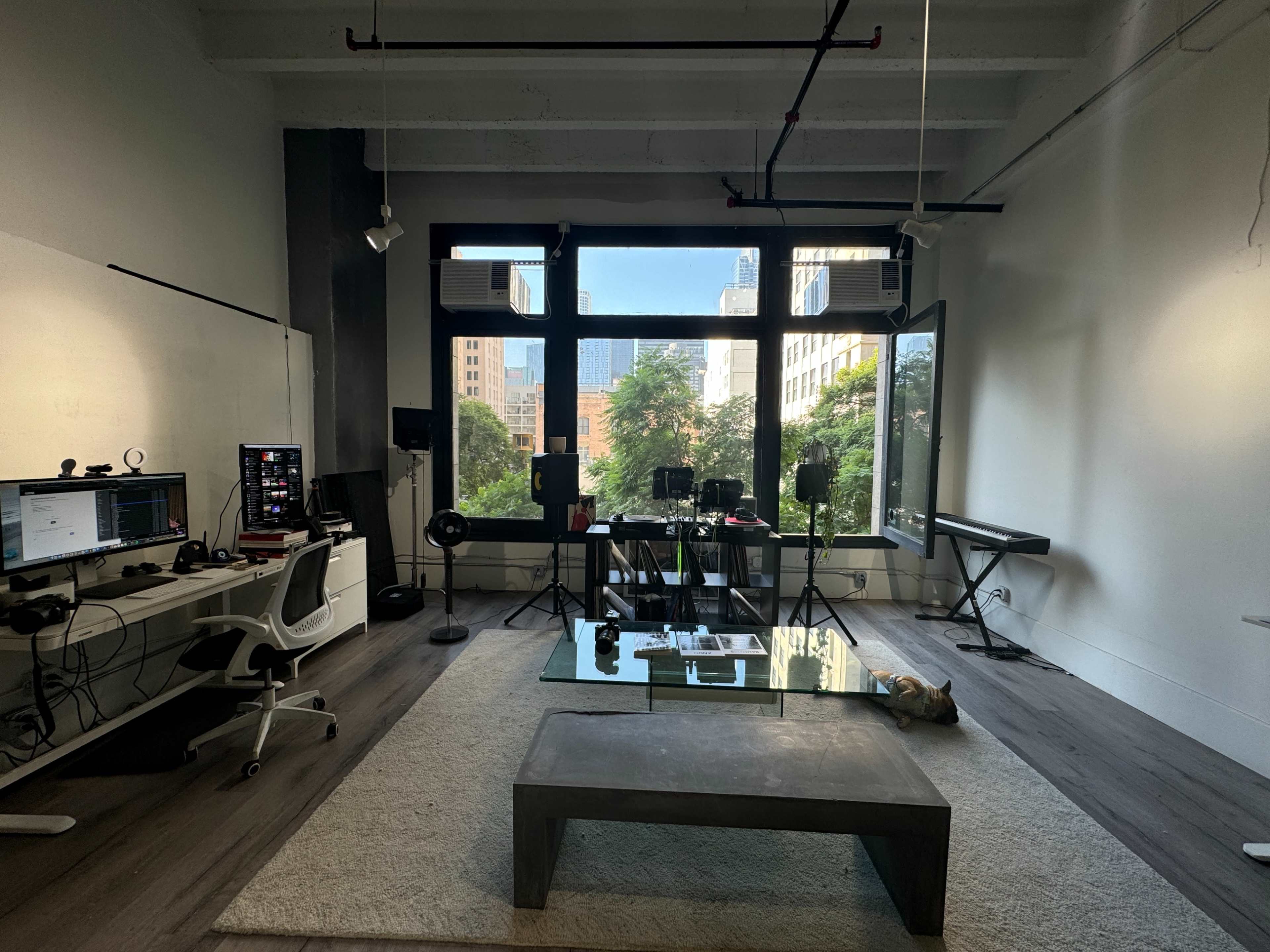The image shows a modern office space with large windows, a desk setup with computers, audio equipment, and a keyboard, all situated on gray wooden flooring.