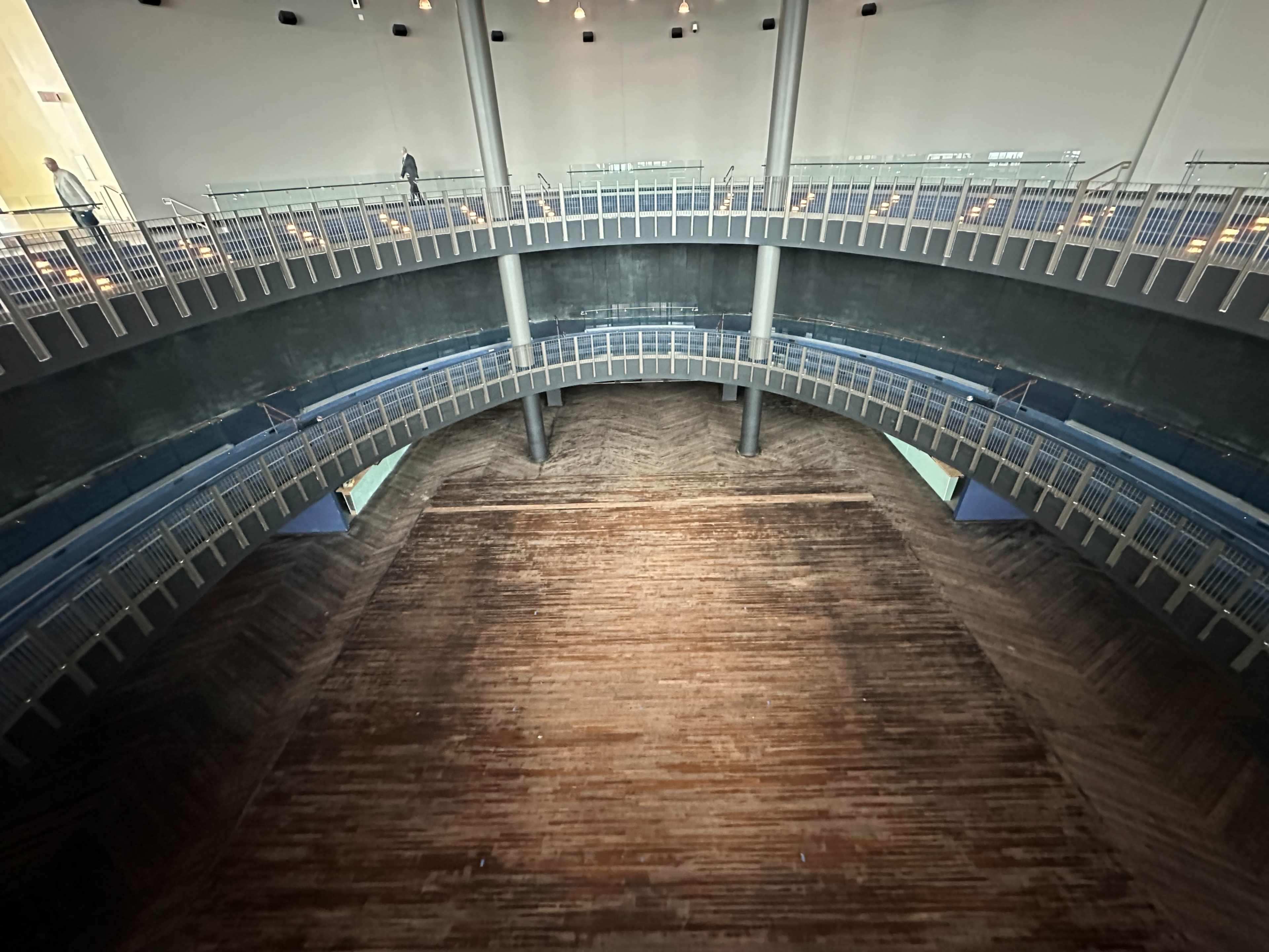 A circular, multi-tiered interior space with a wooden floor and a high ceiling, featuring pathways and a few people walking on the upper levels.