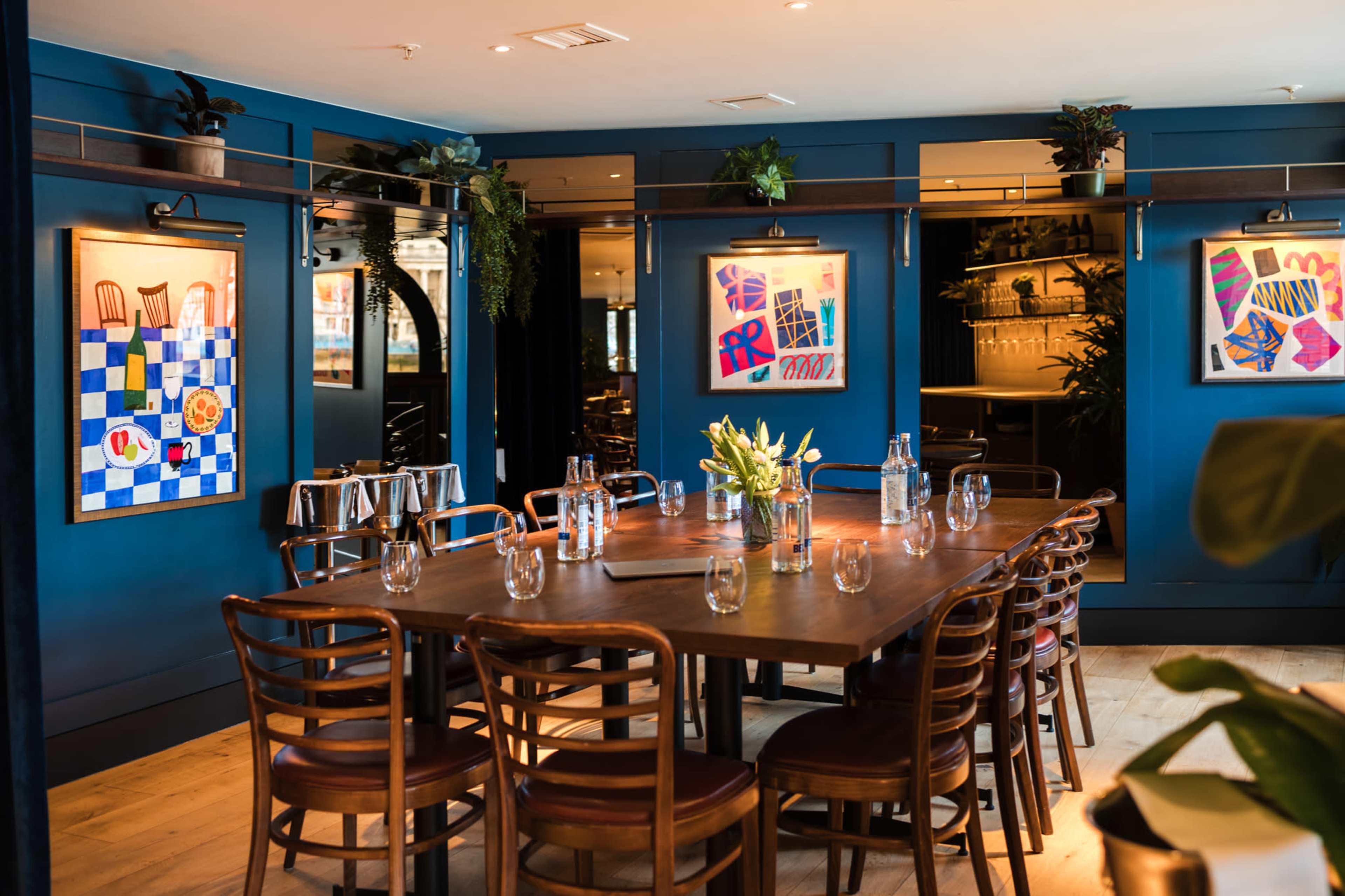 The image shows a well-lit dining room with a large wooden table surrounded by chairs, decorated with vibrant artwork on the blue walls.