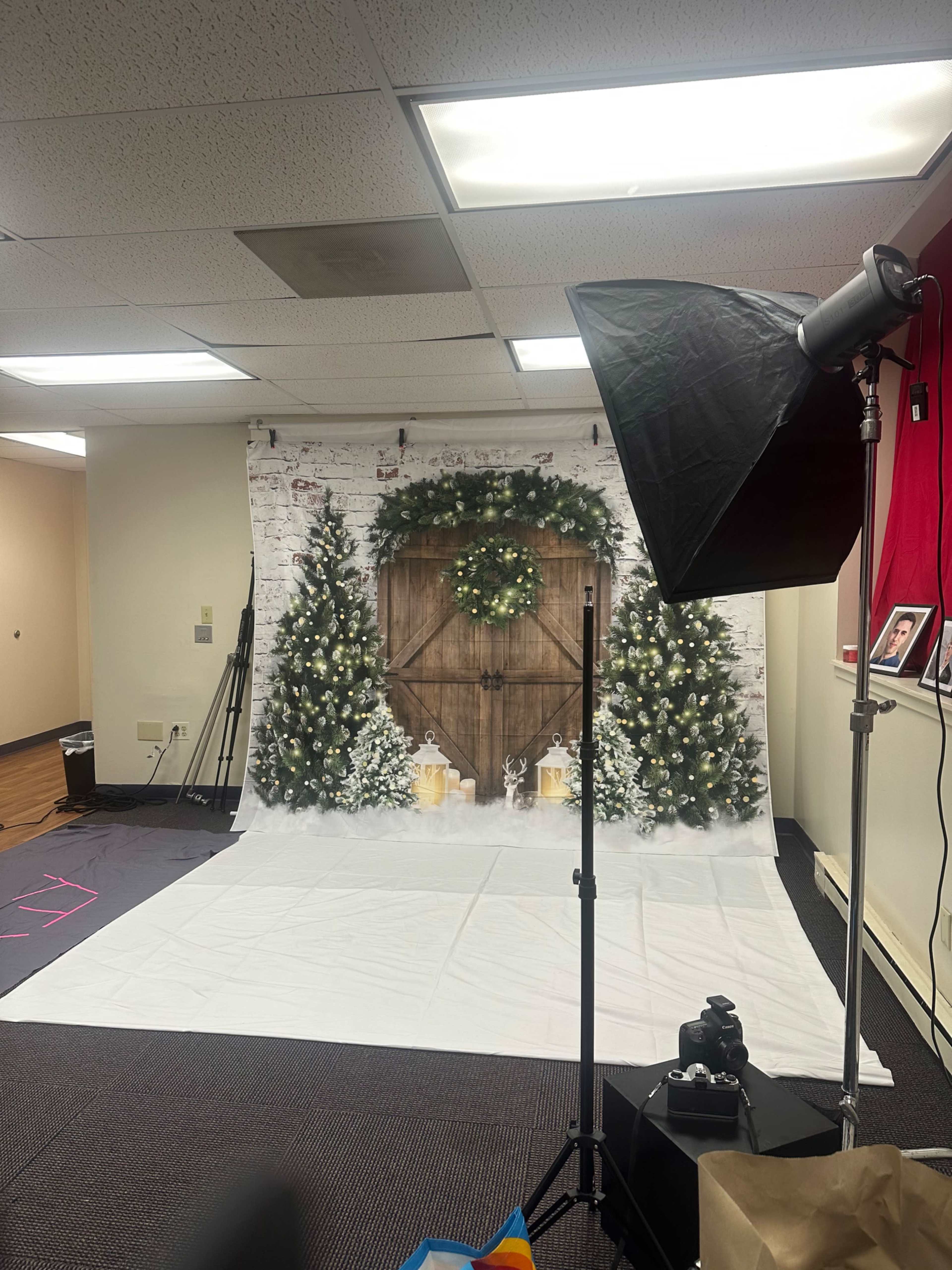 The image shows a photography setup featuring a decorative backdrop with a wooden door adorned with a wreath and flanked by Christmas trees, along with studio lighting equipment.