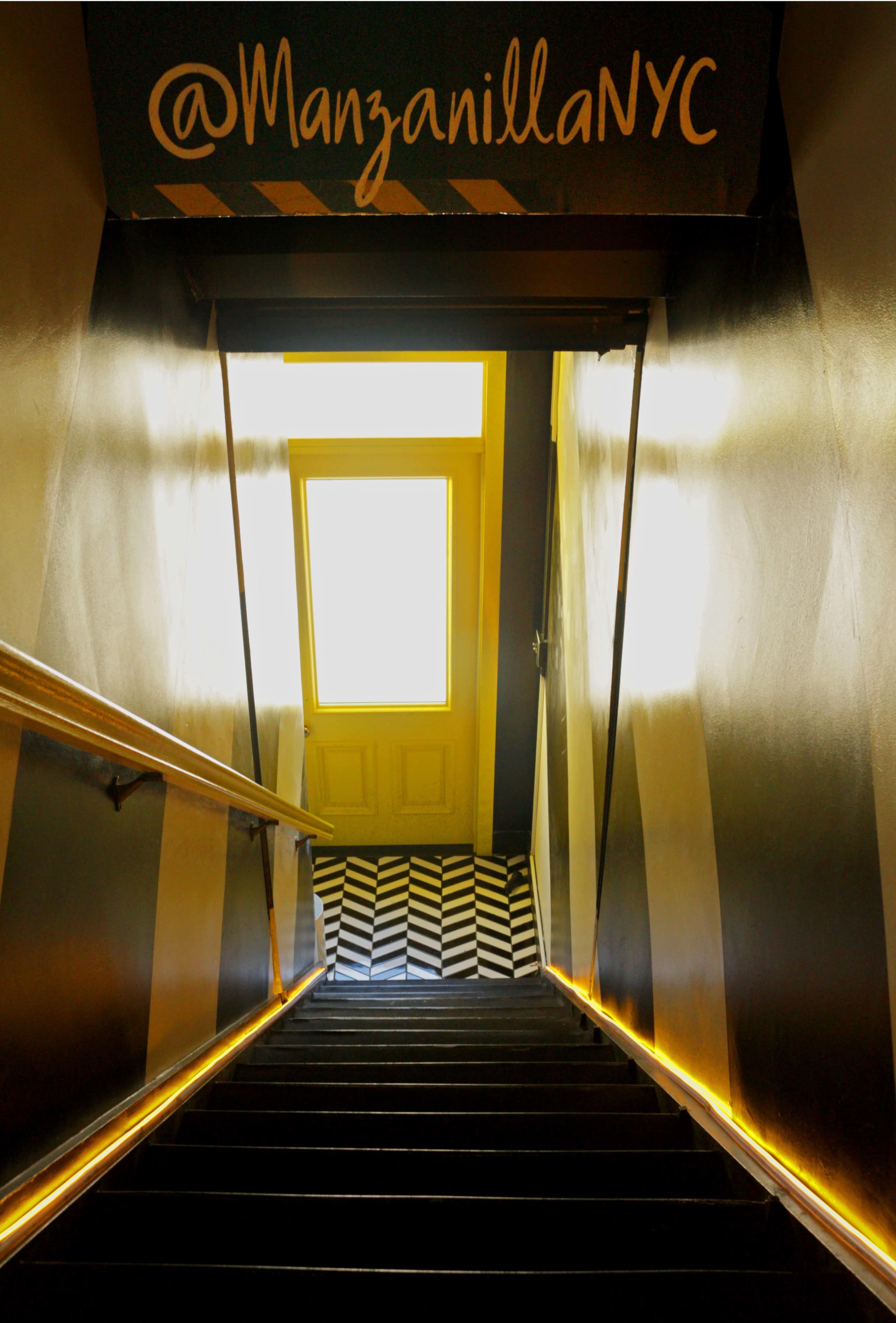 A staircase descends toward a large, brightly lit open door, with patterned black and white flooring visible at the bottom.