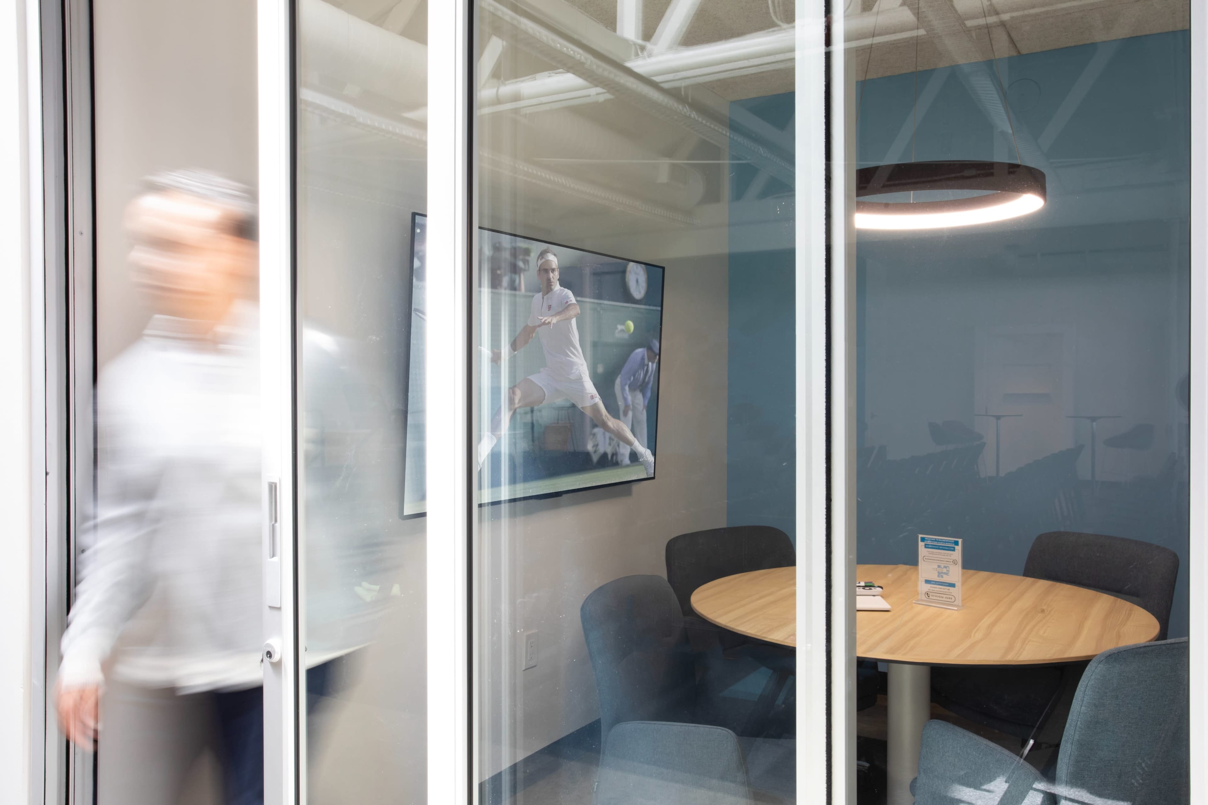 A blurred figure walks past a glass-walled conference room featuring a round table and a large screen displaying a tennis match.
